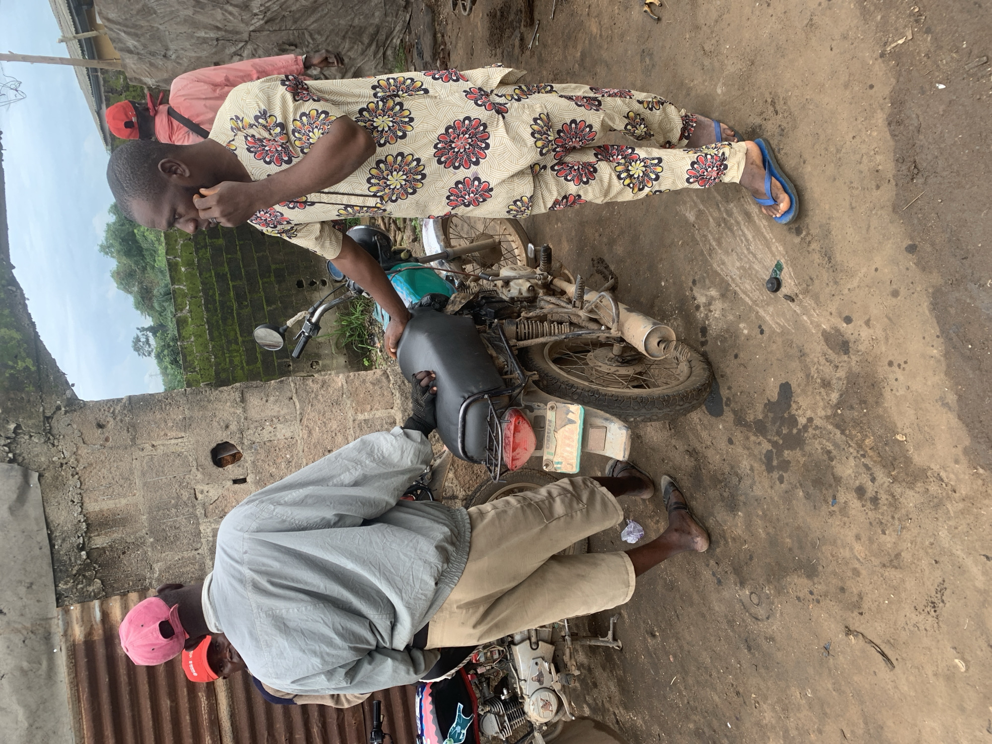 In Shagamu, Nigeria, three men are gathered outdoors near a rough, unfinished concrete structure and a corrugated metal wall. One man, wearing a light gray jacket and tan shorts, stands with his back to the camera, facing a motorcycle. He wears a pink baseball cap and appears to be speaking with another man. To his left, partially obscured, is a man wearing a red baseball cap. The third man, on the right, is dressed in a patterned tunic and trousers with a floral design in red, yellow, and black. He is on the phone and looking to his left, away from the other two men and the motorcycle. The ground is dirt and appears to be muddy in places, with debris scattered around. Two motorcycles are visible, one in the foreground and another partially visible on the left. The scene suggests a casual gathering or a discussion taking place in an outdoor, informal setting.