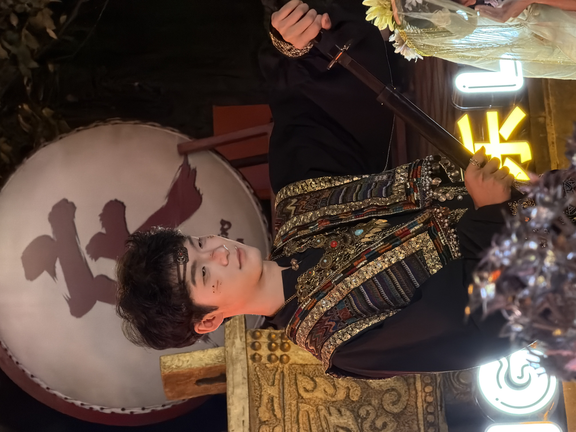 A young East Asian male, dressed in a black garment with an ornate, multi-colored, and gold-trimmed vest, is the central subject of the image. He has dark, wavy hair and is wearing a metallic headband. His face has subtle theatrical makeup, including what appear to be small, dark markings on his cheeks and nose. He is holding a dark, slender object, possibly a sword or staff, horizontally in front of him with his right hand. His left arm is bent at the elbow, with his hand resting on the object.
Behind him, a large, circular drum with a bold, red symbol is visible. To his left, there is a golden, textured architectural element adorned with circular motifs. The background is dark and dimly lit, with hints of foliage and structural elements. In the foreground, there are bright, neon-like signs with Chinese characters. One sign displays a bright yellow character, and to the right, a white character is partially visible. A veil or decorative fabric with gold tassels and flowers hangs over the right side of the frame. The overall setting appears to be an outdoor night scene, possibly a performance or cultural event. The location is identified as Yuzhong District, China.