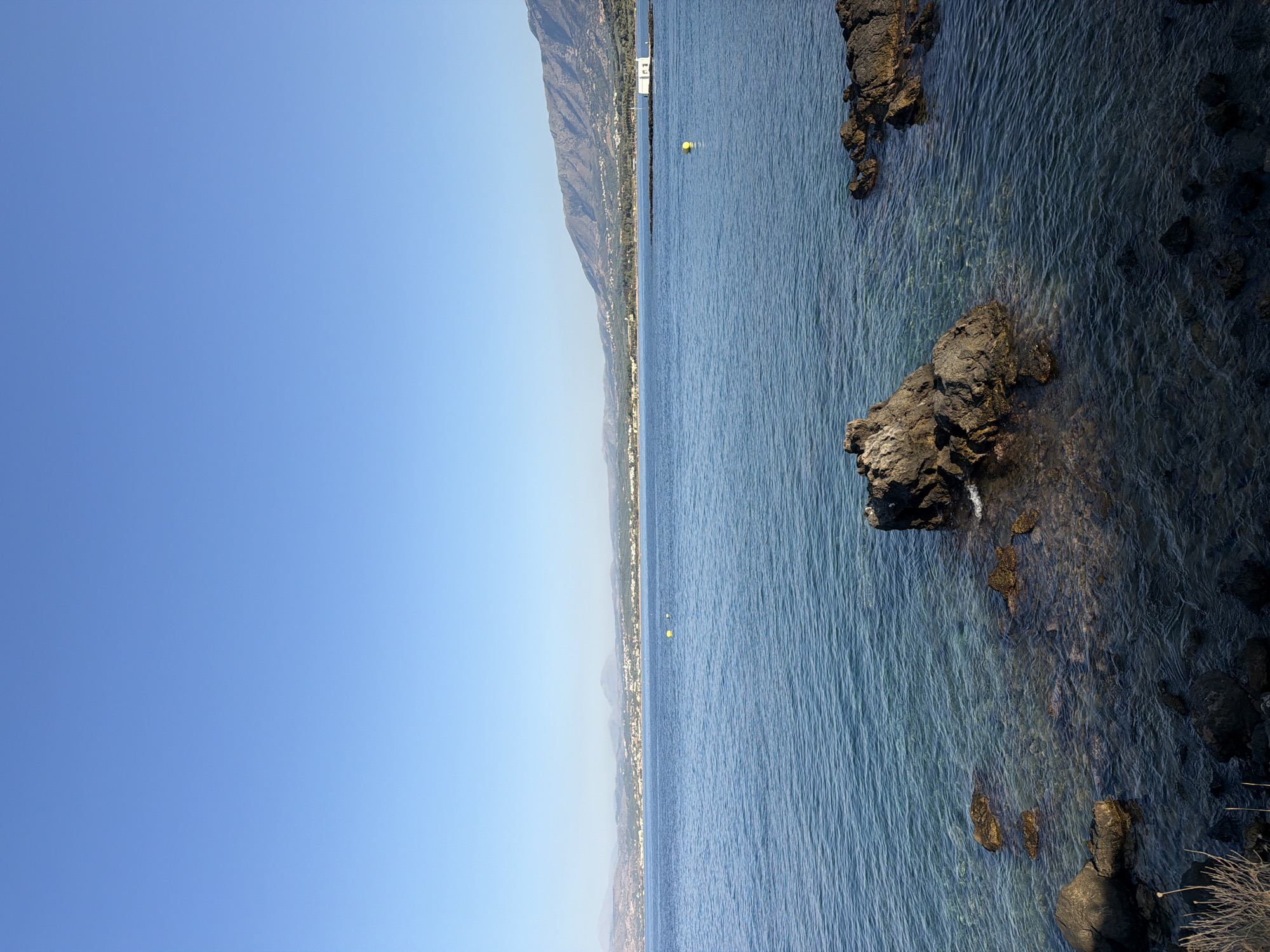 The image shows a serene coastal scene in Georgioupoli, Greece. The foreground is dominated by dark, rocky outcrops in the clear blue water, some partially submerged, revealing the rocky seabed beneath. A larger, prominent rock formation with a textured surface sits in the middle ground.
In the mid-ground, the calm, shimmering blue water stretches towards a distant coastline. The coastline is lined with a low-lying town or village, characterized by a collection of buildings, and further inland, a range of rolling hills and mountains forms the backdrop. The sky is a clear, cloudless expanse of light blue.
Two yellow buoys are visible in the water, one closer to the shore and another further out towards the coastline. A small, white building with a dark roof stands on a small peninsula extending into the sea on the right side of the image. The overall impression is one of tranquility and natural beauty, bathed in clear daylight.