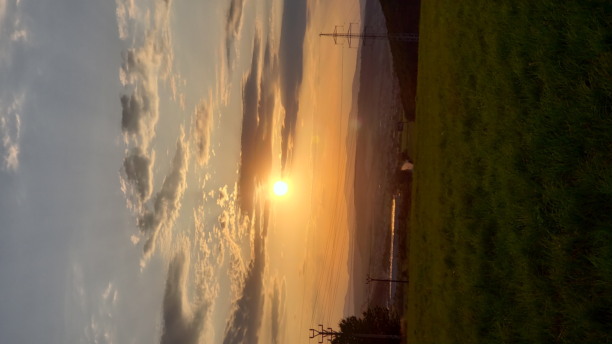 The image depicts a sunset over a landscape near Prešov, Slovakia. The foreground is dominated by a grassy field, rendered in dark green hues due to the low light. In the mid-ground, a valley unfolds, featuring a body of water that reflects the warm, orange glow of the setting sun. A road winds through the valley, leading towards a settlement with buildings and structures. Power lines and utility poles are visible, stretching across the landscape, some silhouetted against the sky. In the distance, rolling hills and mountains form a hazy, layered backdrop. The sky itself is a dramatic canvas of oranges, yellows, and soft blues, with scattered clouds tinged by the sun's rays. The sun, a bright, luminous orb, is positioned in the upper left quadrant of the image, casting a warm light over the entire scene. The overall impression is one of a serene, natural landscape bathed in the warm light of a setting sun.