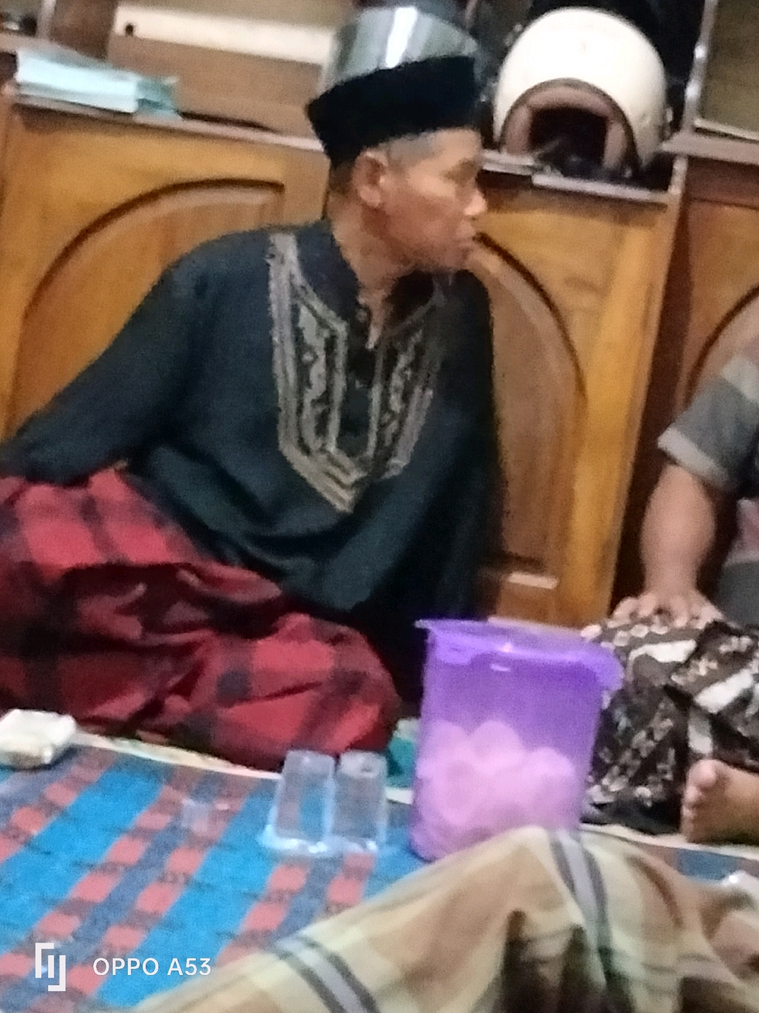 The image shows an indoor scene, likely a room in a home. The primary subject is a man wearing a black peci (a type of cap common in Indonesia) and a dark blue or black robe with intricate gold embroidery on the chest. He is also wearing a red and black checkered sarong wrapped around his waist. He is seated, facing to the right of the frame, and appears to be looking at something or someone outside the frame.
In the background, there is wooden furniture, possibly a cabinet or dresser, with a white helmet resting on top of it. To the right of the man, a portion of another person's arm and torso is visible, wearing a striped shirt. In the foreground, on a mat with blue and red checkered stripes, there are several objects, including a translucent purple container filled with pink spherical items (possibly cotton candy or small balls), two small translucent cups, and a white rectangular object. The lighting suggests an indoor setting with ambient light. There is no specific indication of the city of Trenggalek, Indonesia within the image itself, and its identification would require external information.