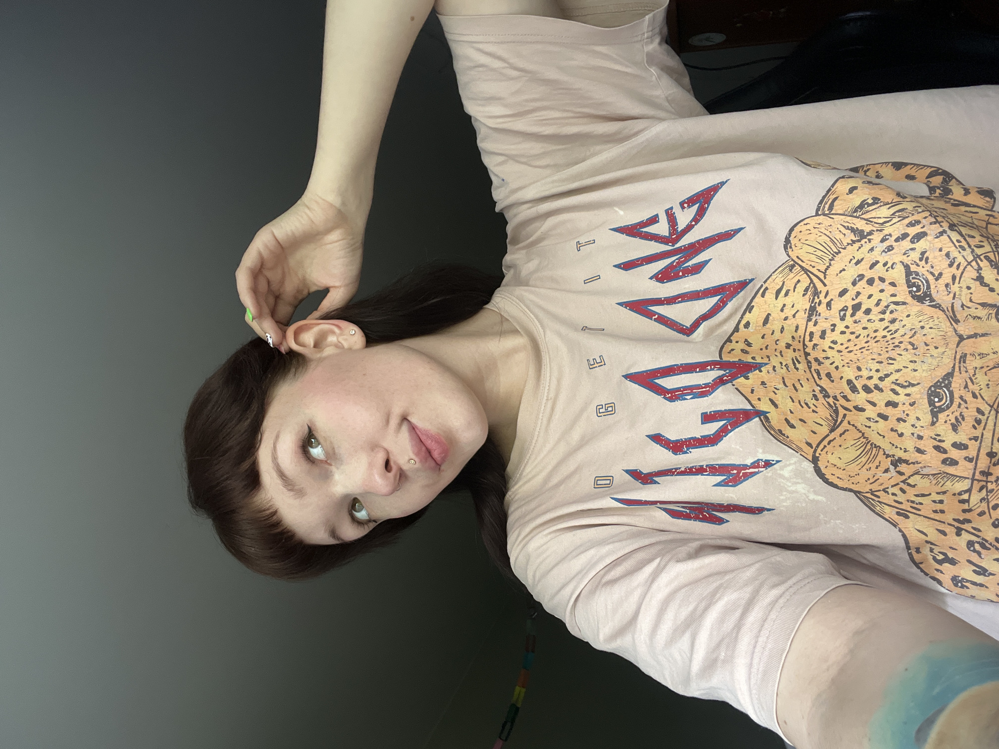 A young woman with brown hair and green eyes is looking at the camera. She is wearing a light pink T-shirt with a graphic design that includes a leopard's head. Her left hand is near her ear. The background is a grey wall. It is unlikely that the location in Chaikovsky, Russia can be determined.