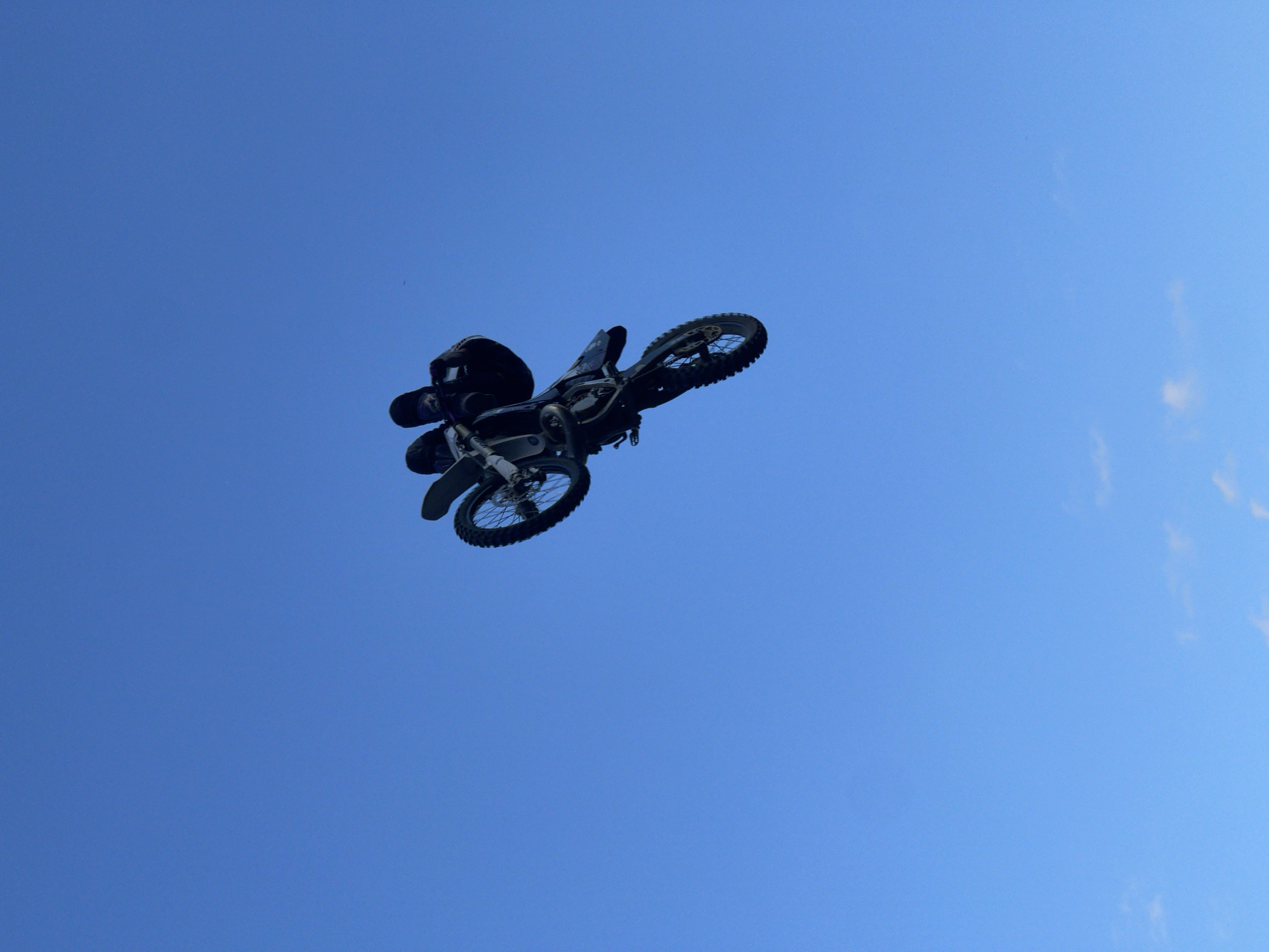A person is performing a stunt on a dirt bike in mid-air against a clear blue sky. The dirt bike is upside down, and the rider is in a tucked position, appearing to be mid-jump or trick. The background is an expansive, cloudless blue sky, suggesting an outdoor event or location. The specific location within Strathroy-Caradoc, Canada, is not discernible from the image itself.