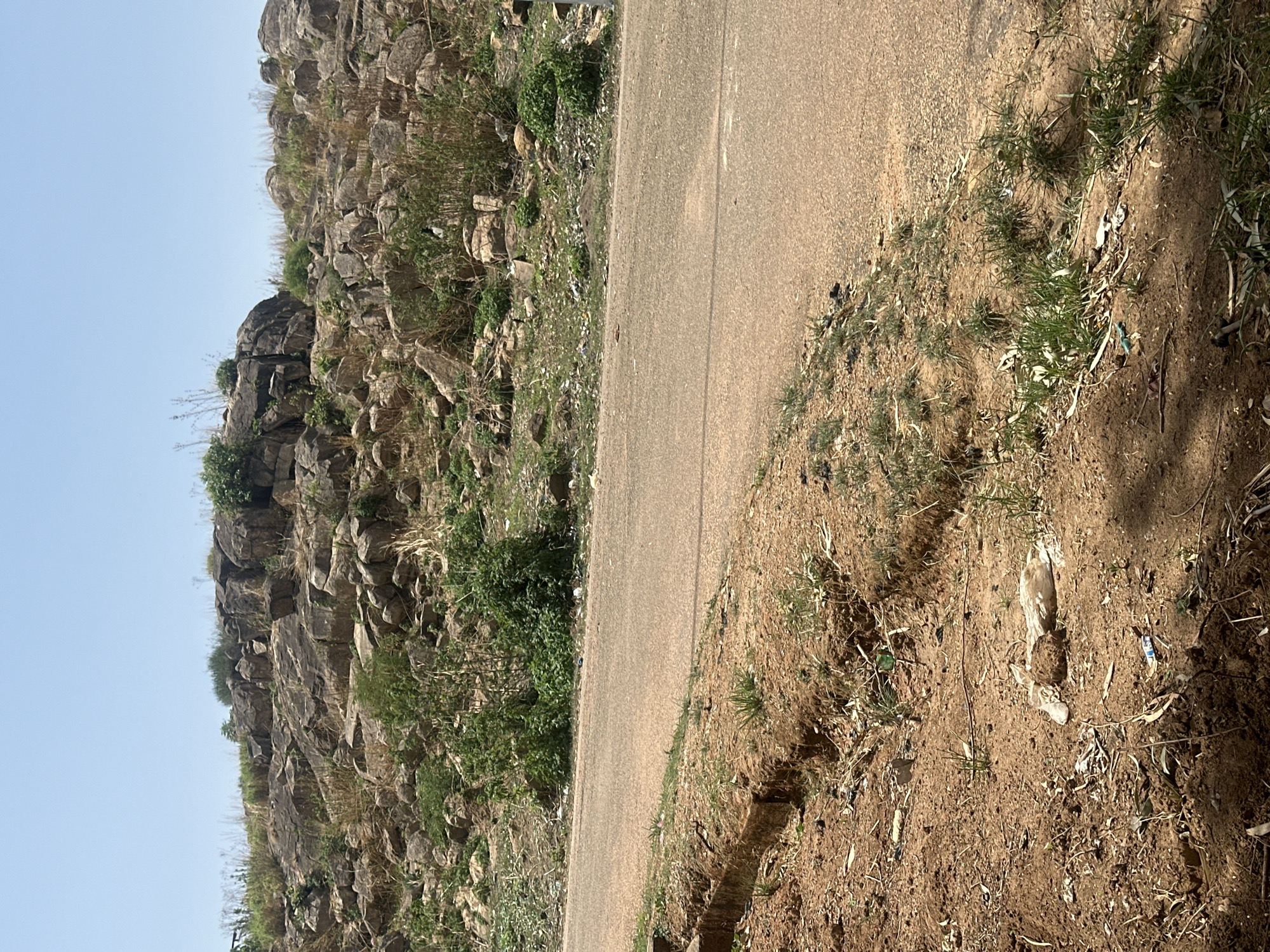 The image depicts a roadside scene in Jos, Nigeria. A paved road runs diagonally across the frame, bordered on one side by a rocky hillside covered in sparse vegetation. The opposite side of the road is characterized by a dirt embankment with patchy grass and scattered debris, including plastic waste. The sky is a pale blue. The overall impression is one of a dry, rural or semi-rural environment. No people, subjects, actions, or interactions are visible in the image.