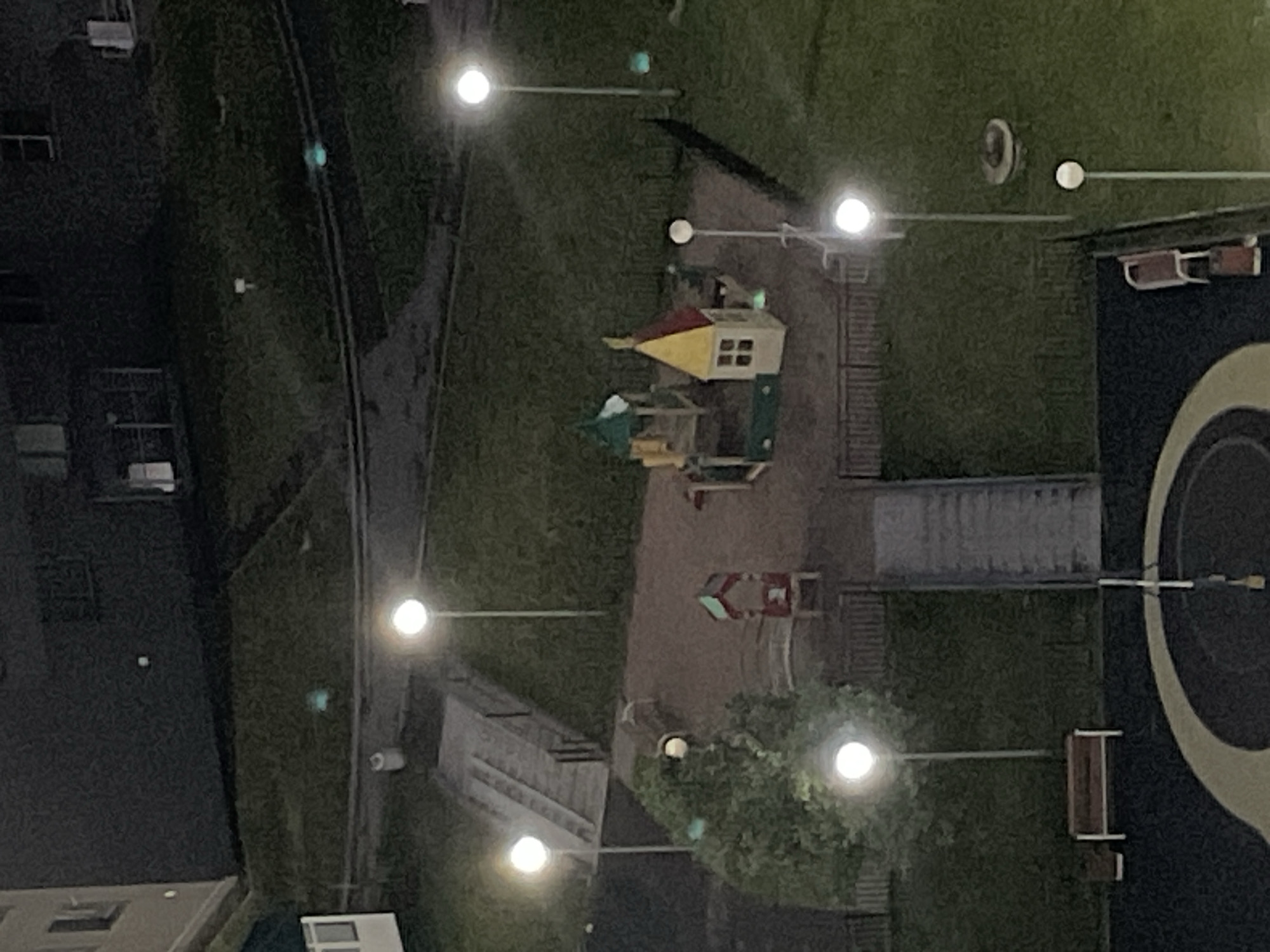 The image shows an outdoor courtyard at night, likely in the city of Sinyavino, Russia, given the context. The scene features a children's playground with a small playhouse and a climbing structure. Several lampposts are illuminated, casting light on the grassy area and the playground equipment. A set of outdoor stairs leads up to a pathway. There is also a bench visible. The surrounding environment appears to be a residential area with apartment buildings in the background. The ground surface is a mix of grass and paved areas. No people are visible in the image.