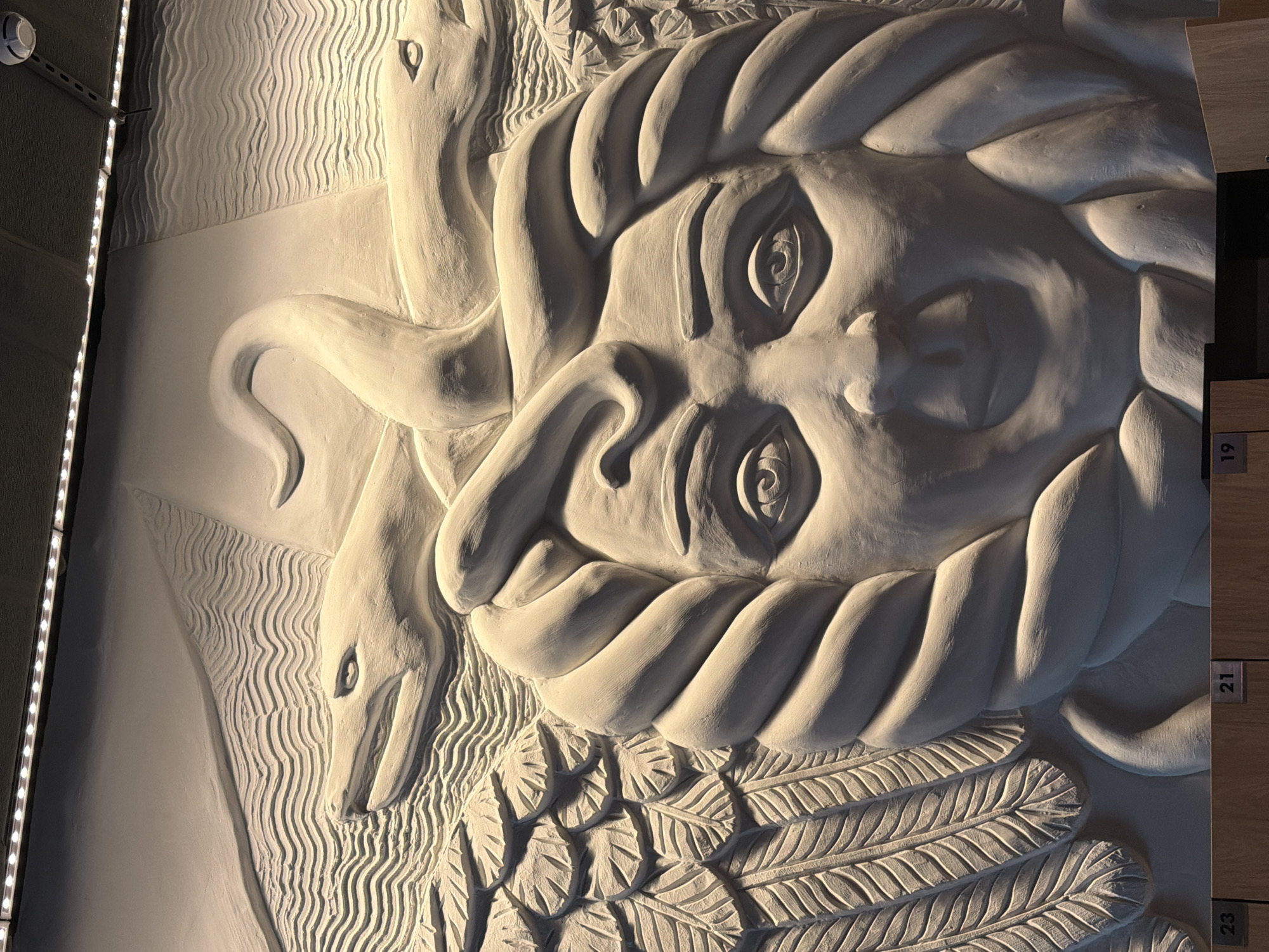This is a close-up, low-angle shot of a large, light-colored bas-relief sculpture depicting a stylized face with wings and two serpent-like creatures. The sculpture is illuminated by overhead lighting, creating strong shadows that highlight the texture and form of the artwork. The face is rendered with wide, watchful eyes, a prominent nose, and pursed lips. Intricate feather patterns form the wings on either side of the face. Two serpentine forms intertwine above the head of the face, with one appearing to coil around the other. The background of the sculpture features wavy, undulating lines, possibly representing water or wind. The lower portion of the image shows the edge of a wooden structure with numerical labels "23," "21," and "19," suggesting this sculpture is part of an exhibition or installation. The style of the sculpture, with its mythological and symbolic elements, could be found in various cultural institutions in Saint Petersburg, Russia, such as a museum, gallery, or public space dedicated to art.