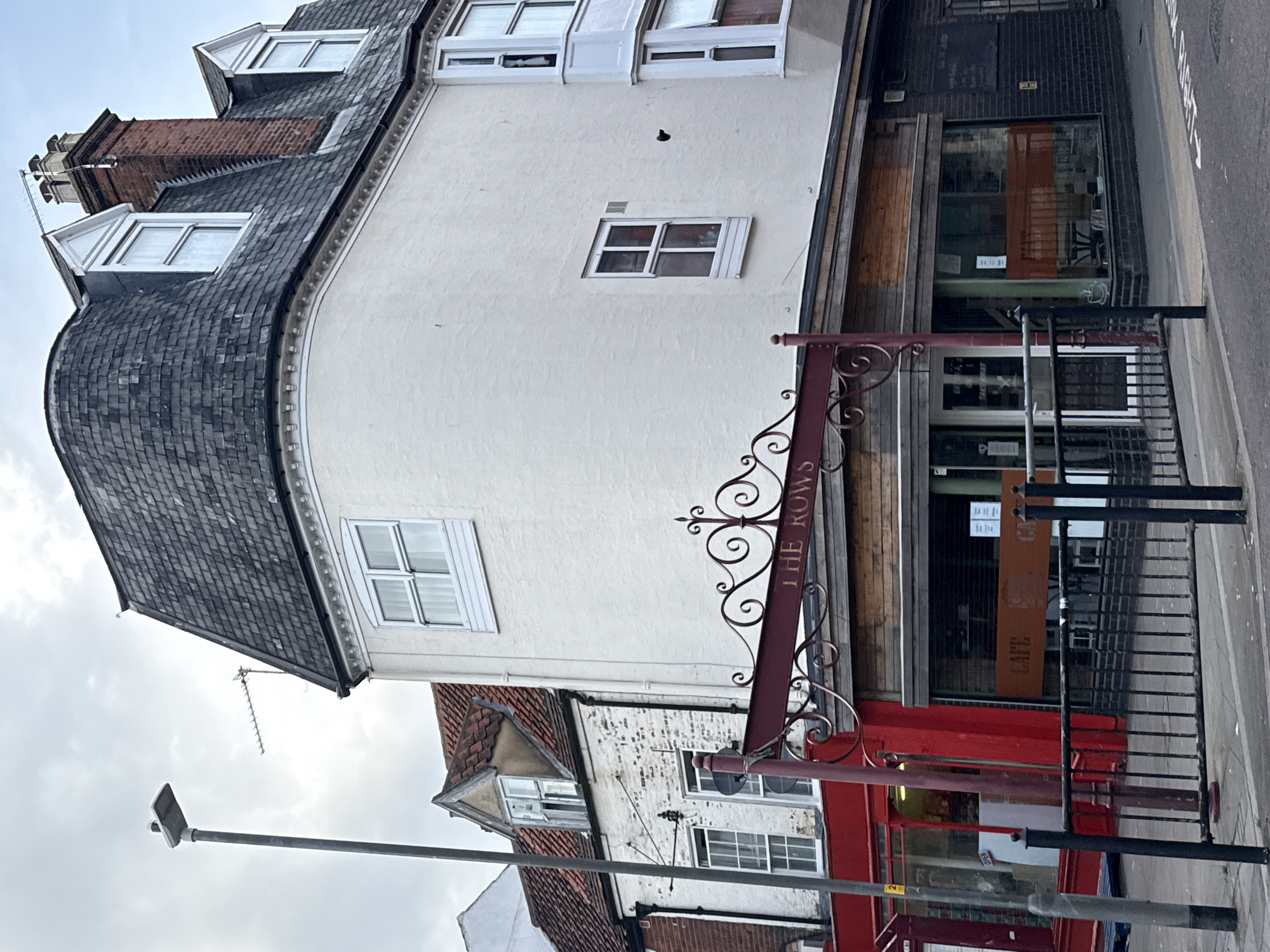 The image depicts a section of a street in Great Yarmouth, United Kingdom, featuring a red sign reading "THE ROWS." The sign is attached to a building with a white painted facade and multiple windows. The architecture is multi-storied, with a curved roofline. The building appears to house shops or businesses on the ground floor, indicated by storefront windows and wood paneling. Adjacent buildings display varied architectural styles and colors. A black metal railing runs along the sidewalk. A street lamp is visible. The sky is overcast.