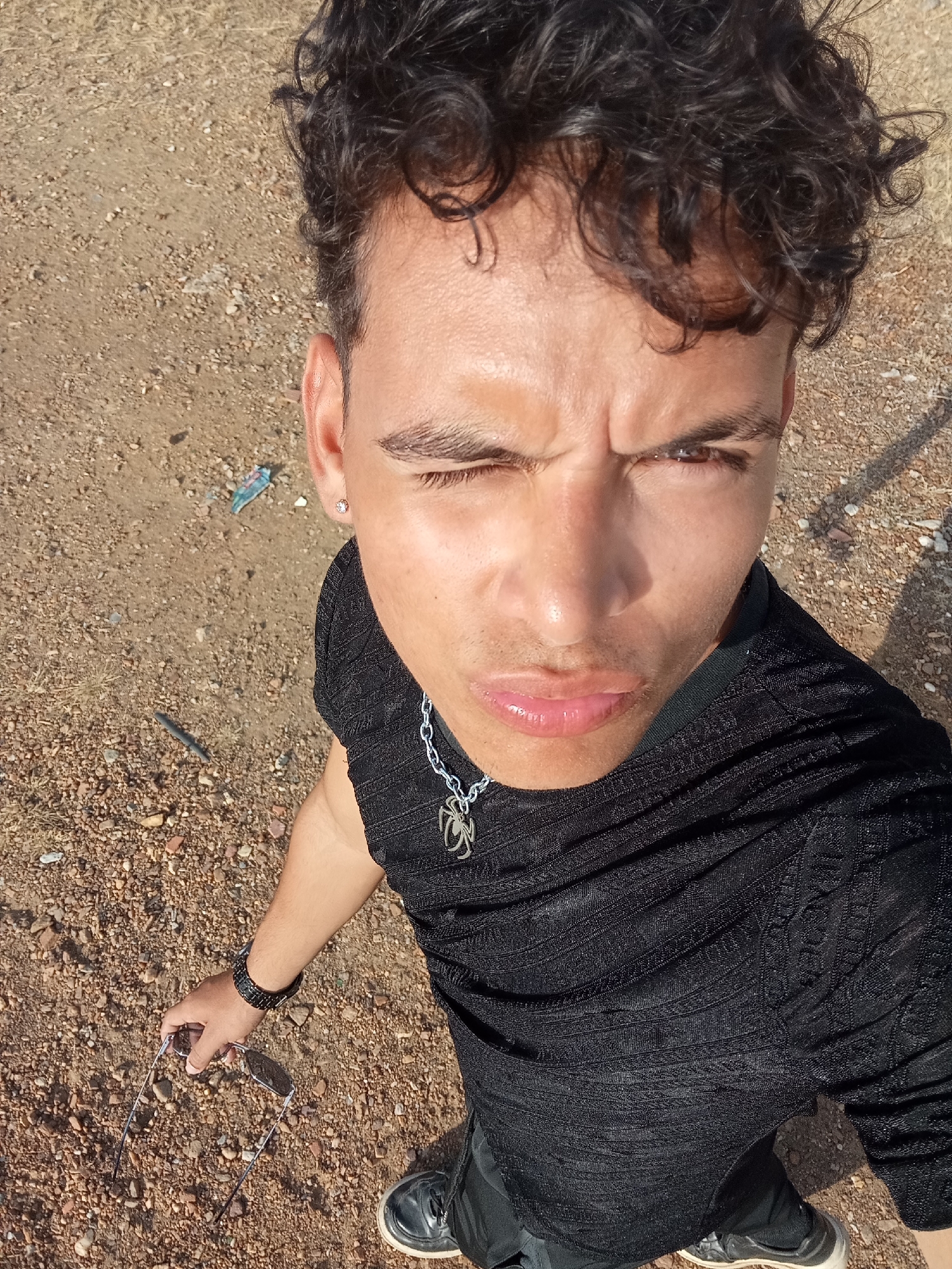 This high-angle selfie captures a young man outdoors, likely in Juan Griego, Venezuela, under bright daylight. He looks directly up at the camera with an intense expression, characterized by a squinted right eye, furrowed forehead, and slightly pursed lips. He has dark, curly hair.
The man is wearing a black, textured or patterned short-sleeved shirt and a silver chain necklace adorned with a distinctive spider-shaped pendant. A small, clear stud earring is visible in his left ear. On his left wrist, he wears a dark-colored watch, and in his left hand, he holds a pair of open sunglasses.
The background is a dry, earthy-toned ground covered with light brown and reddish-brown gravel and dirt. Small pieces of debris, including a blue wrapper and a dark, stick-like object, are scattered on the ground. The strong sunlight casts prominent shadows, indicating a clear, sunny day. No text is visible in the image.