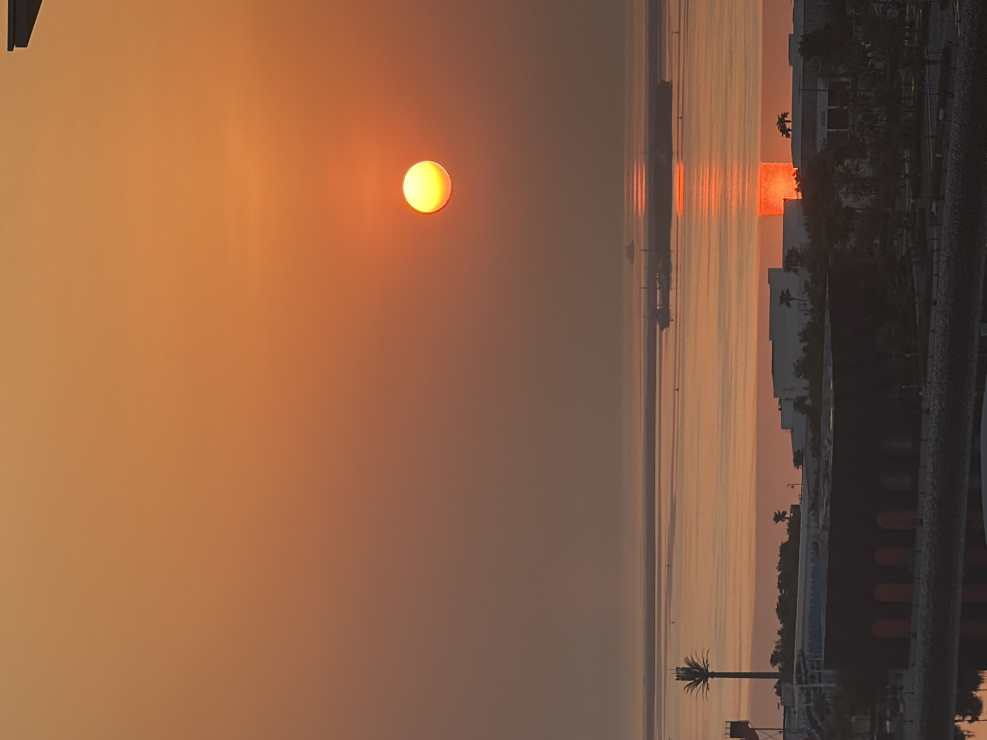 The image shows a sunset over the city of Lusail, Qatar. The sun, a bright orange orb, is setting on the horizon, casting a warm glow over the scene. The sky is a gradient of orange and yellow, with a few wispy clouds. In the foreground, there are several buildings, silhouetted against the sunset. One building is particularly prominent, with a dark, rectangular shape and arched doorways. There is also a tall palm tree, its silhouette stark against the vibrant sky. In the distance, a large ship is visible on the water, its reflection shimmering on the surface. The water itself appears calm and reflects the colors of the sunset. The overall impression is one of serenity and natural beauty, a tranquil end to the day in Lusail.