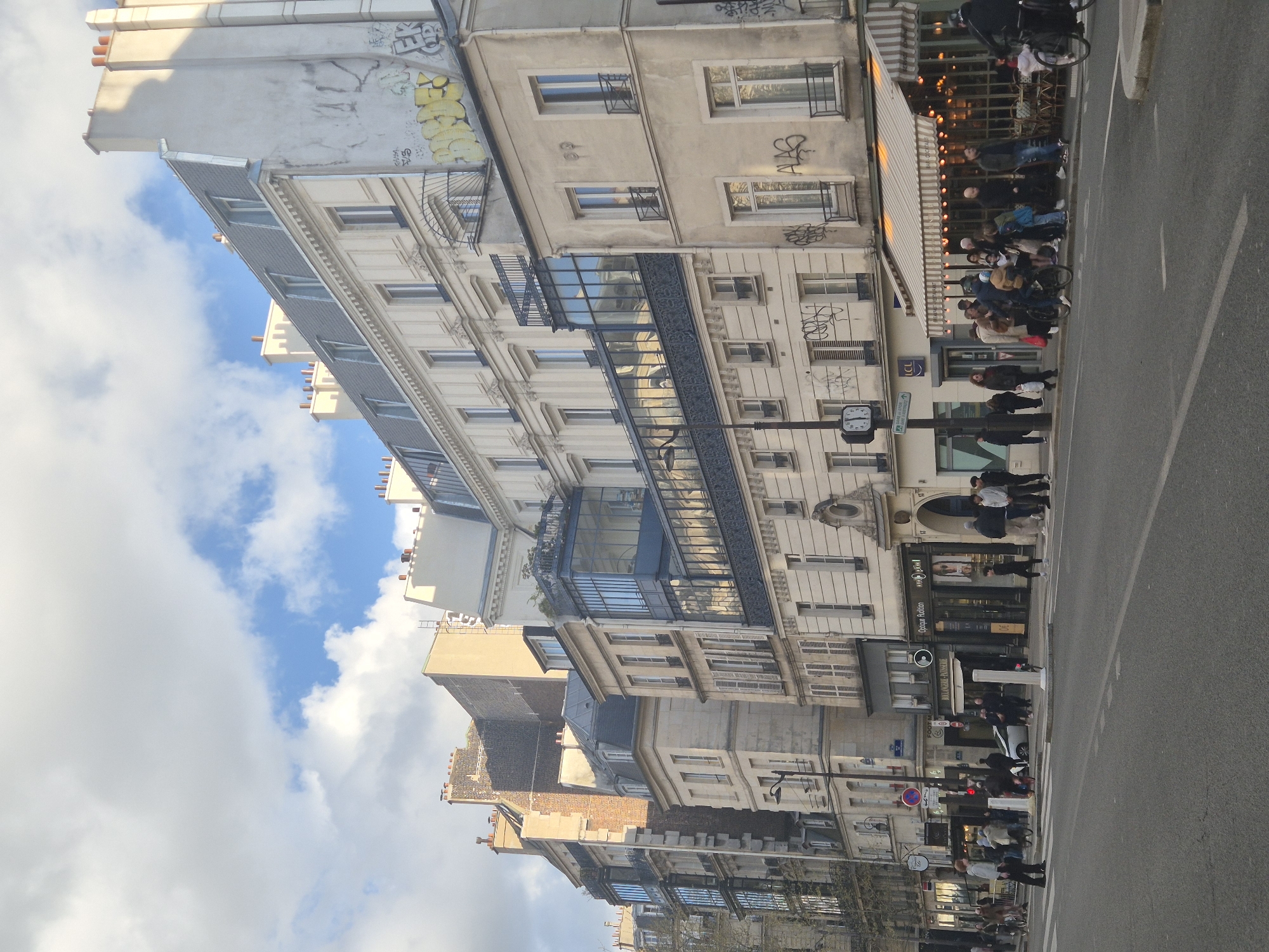 This image depicts a street scene in Paris, France. Several buildings line the street, showing classic Parisian architecture with multiple stories, ornate facades, and windows. Along the sidewalk, there are people walking and standing, suggesting a lively urban atmosphere. A restaurant appears to be open with outdoor seating, indicating activity and interaction. A street sign with clock is visible, adding to the authenticity of a typical Parisian street view. The sky is cloudy, with patches of blue, providing light to the scene. The presence of a road and sidewalk further clarifies the scene's urban environment.
