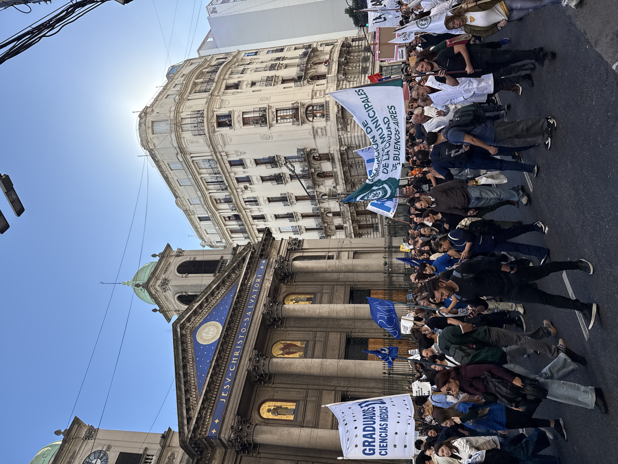 A crowd of people marches down a city street in Buenos Aires, Argentina. A baroque-style building with a green dome is on the left; it is the Church of El Salvador in Buenos Aires. The marchers carry signs and banners. A light-colored building is on the right side of the street. Power lines are visible at the top left. It is daytime, and the sun is shining.