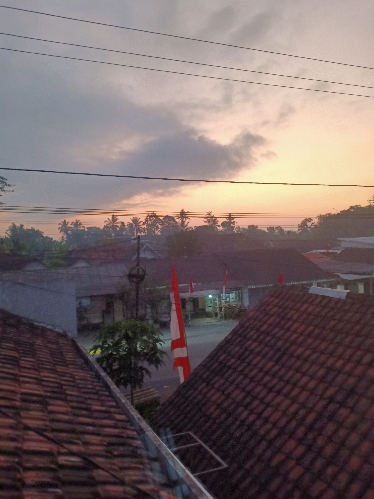 The image depicts a view from a rooftop overlooking a village scene in Jarit, Indonesia, during what appears to be sunrise or sunset. The sky is a gradient of soft yellows and oranges, with some grey clouds present. Several power lines run horizontally across the upper portion of the frame. Below the sky, a line of trees, including palm trees, forms a silhouette against the horizon. The foreground is dominated by the red-tiled roofs of houses. In the mid-ground, more houses with red-tiled roofs are visible, some with visible doorways or windows. There are indications of small shops or businesses with awnings. A flagpole with the Indonesian flag, red and white, is prominently displayed on the right side of the mid-ground. A street or pathway is visible between the buildings. A small tree is situated in the mid-ground. The overall atmosphere is serene and rural.
