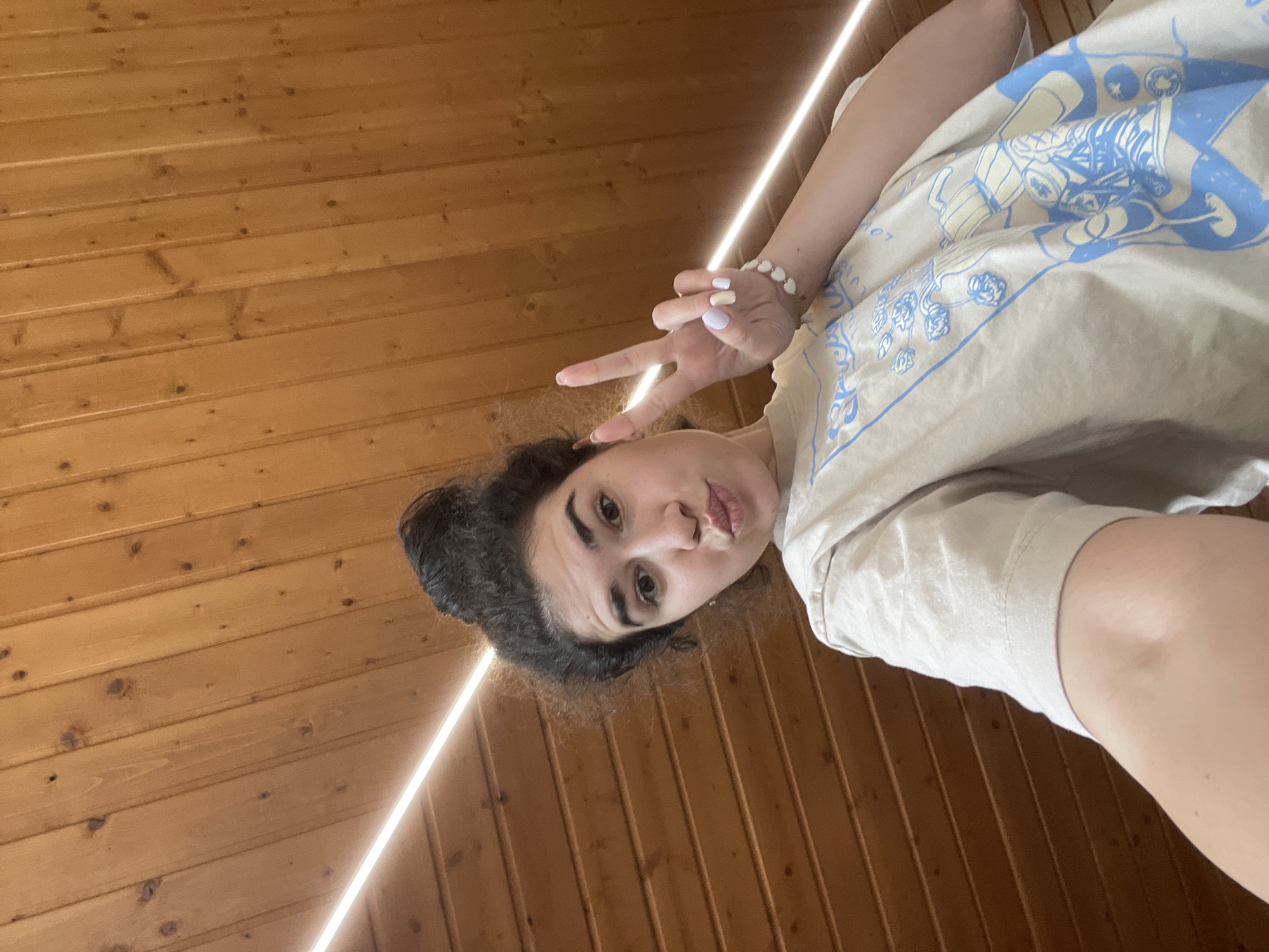 A person, identified as female, is shown in a selfie-style photograph. She has dark curly hair pulled into a bun. She is wearing a light-colored t-shirt with a blue graphic design. Her fingernails are painted in light colors. She is making a peace sign with her fingers. The background appears to be a wood-paneled wall with a light fixture. The location is undetermined, but is presumed to be indoors. No city details are present.