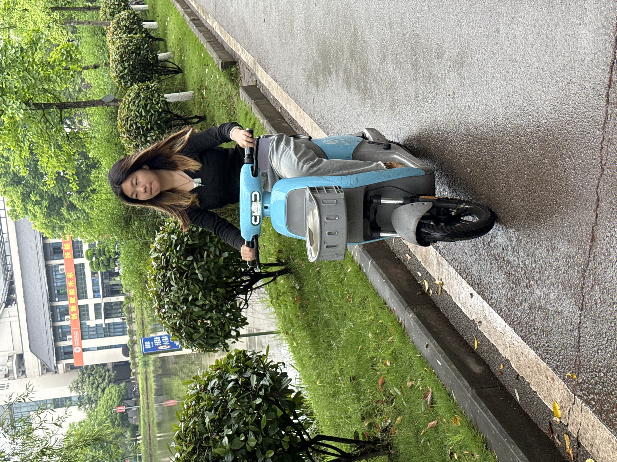 A young woman is riding a light-blue electric scooter on a wet asphalt road in an outdoor setting. The scooter is positioned on the right side of the road, which has a white painted line separating it from a grassy area with trees and shrubs. On the left side of the road, there is a body of water. In the background, there is a multi-story building with Chinese writing on a banner. The photo was taken in the Jiangjin District of Chongqing, China.