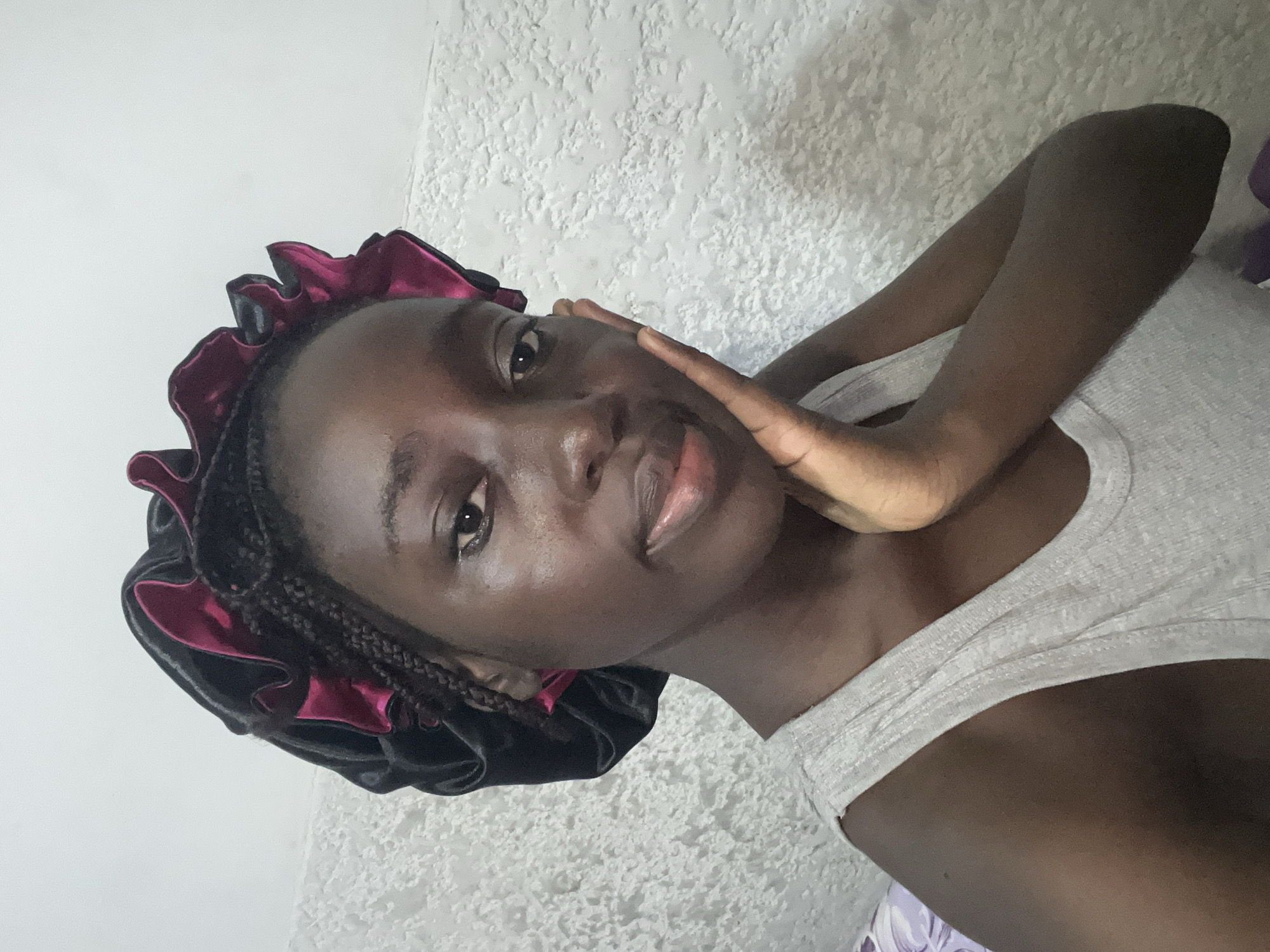 A young woman with dark skin is the focus of this image, which is oriented vertically and appears to be a selfie. She is wearing a light gray tank top and a black hair bonnet with maroon ruffles, and her hair is braided. The woman's right hand is gently touching her cheek. Her gaze is directed toward the camera. The background consists of a white wall with a textured surface, and a glimpse of a light purple floral pattern is visible at the bottom. No specific details point to the location being in Abuja, Nigeria. The scene seems to be indoors, potentially in a bedroom or living space.