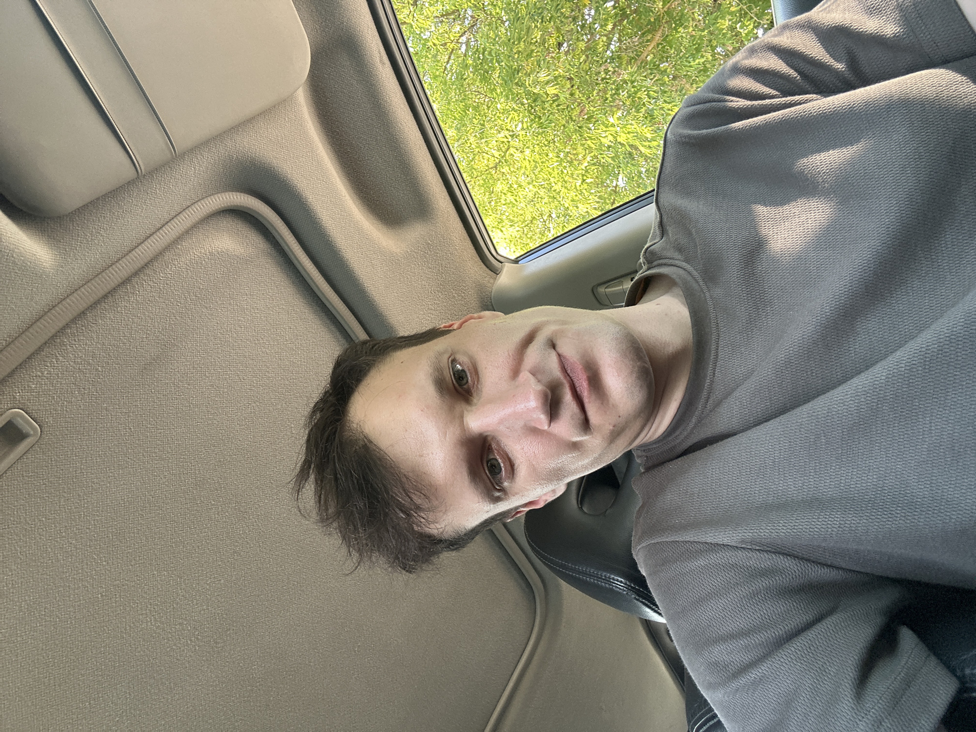 A man is seen from the chest up, taking a selfie from inside a car. He has short, messy brown hair and is wearing a grey t-shirt. The interior of the car is visible, including the ceiling and a sun visor. Through the open window to the right, bright green foliage from trees is visible, suggesting an outdoor setting possibly within a park or residential area of Makiv, Ukraine.