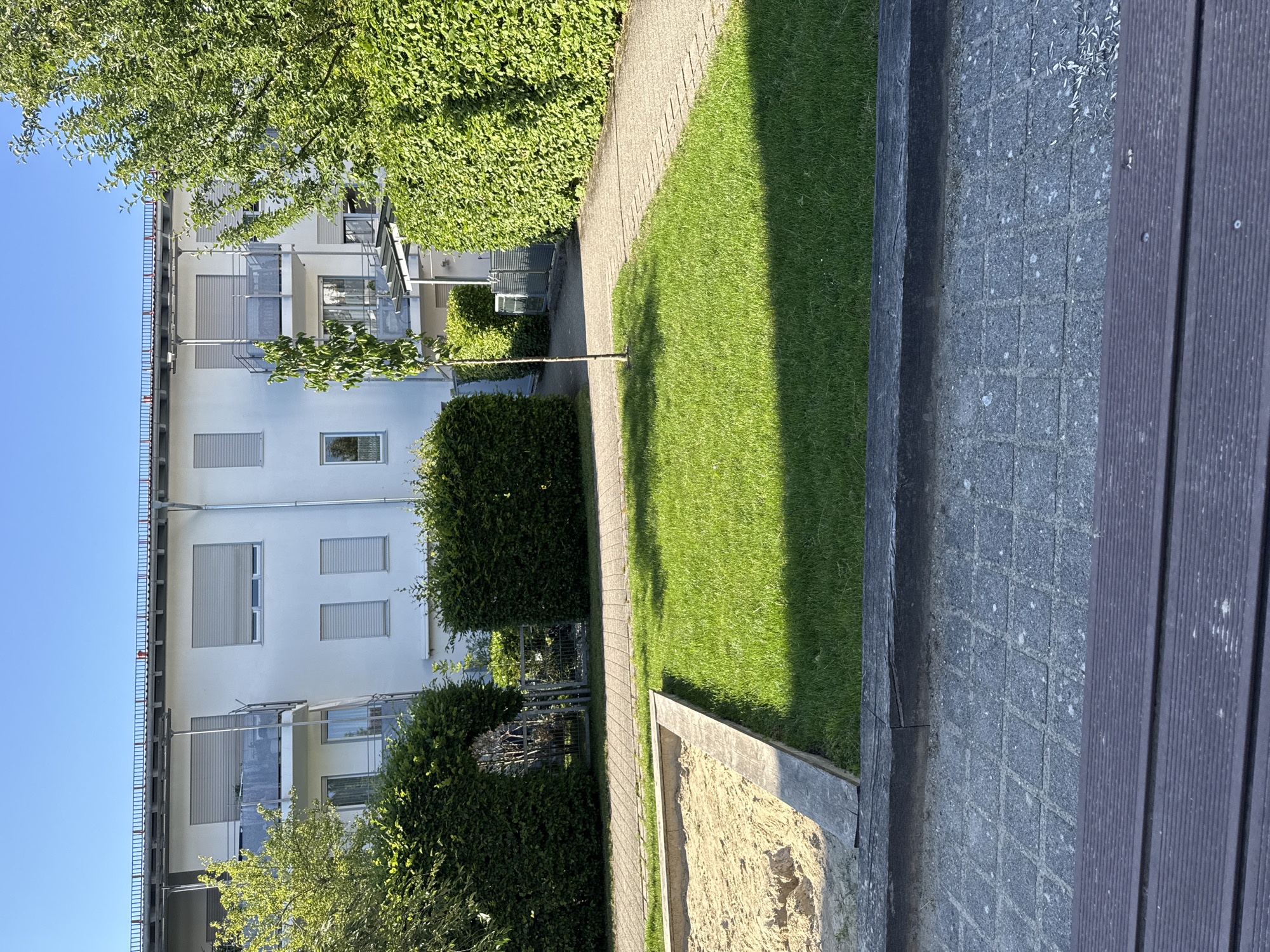 This media file depicts a residential building with a well-maintained outdoor area. The building is modern, with a white facade, multiple balconies, and grey roller shutters on the windows. It appears to be a multi-story apartment complex. Lush green hedges and trees surround the building, creating a pleasant and private atmosphere.
Adjacent to the building, a paved walkway runs alongside a vibrant green lawn. The lawn is neatly mowed and casts a long shadow, indicating that the sun is positioned at an angle, suggesting either morning or late afternoon. A sandbox with a wooden border is visible at the edge of the lawn, hinting at a space for children's recreation. The ground surrounding the sandbox is a mosaic of grey tiles. A dark wooden bench or seating area is present to the right of the frame, possibly part of a patio or outdoor seating arrangement.
The sky is a clear, bright blue, suggesting fair weather. The scene is tranquil and peaceful, evoking a sense of suburban or urban green space. The overall impression is one of a comfortable and modern living environment. No people are visible in the image, and there is no visible text. The context of Höhenkirchen-Siegertsbrunn, Germany, suggests a European setting.