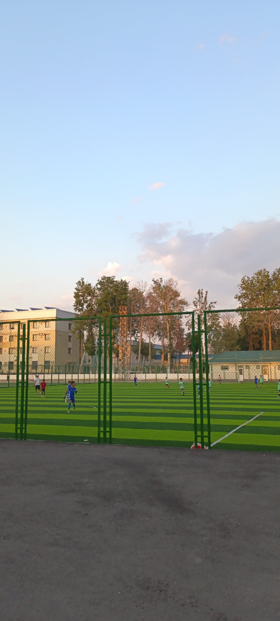 The image depicts an outdoor artificial turf soccer field with a group of children playing. The field is green with white markings and surrounded by a green metal fence. In the background, there are buildings, trees, and a cloudy sky. The time of day appears to be late afternoon or early evening, given the soft lighting and the angle of the sun. The weather seems pleasant, with a clear sky and some scattered clouds.
Several children are visible on the field, engaged in a soccer game. One child in a blue uniform is dribbling a ball, while others are running and positioning themselves. The children appear to be actively participating in the game, suggesting a youth soccer practice or match.
In the distance, a multi-story building with solar panels on its roof is visible. Other structures, including a smaller building with a green roof and windows, are also present. The presence of these buildings and the style of the structures might suggest an urban or suburban setting. The trees in the background are lush and green, indicating a temperate climate.
The overall scene is one of an active and healthy outdoor activity. The focus is on the children's engagement with the sport, set against a backdrop of a developed urban environment. There is no visible text in the image.