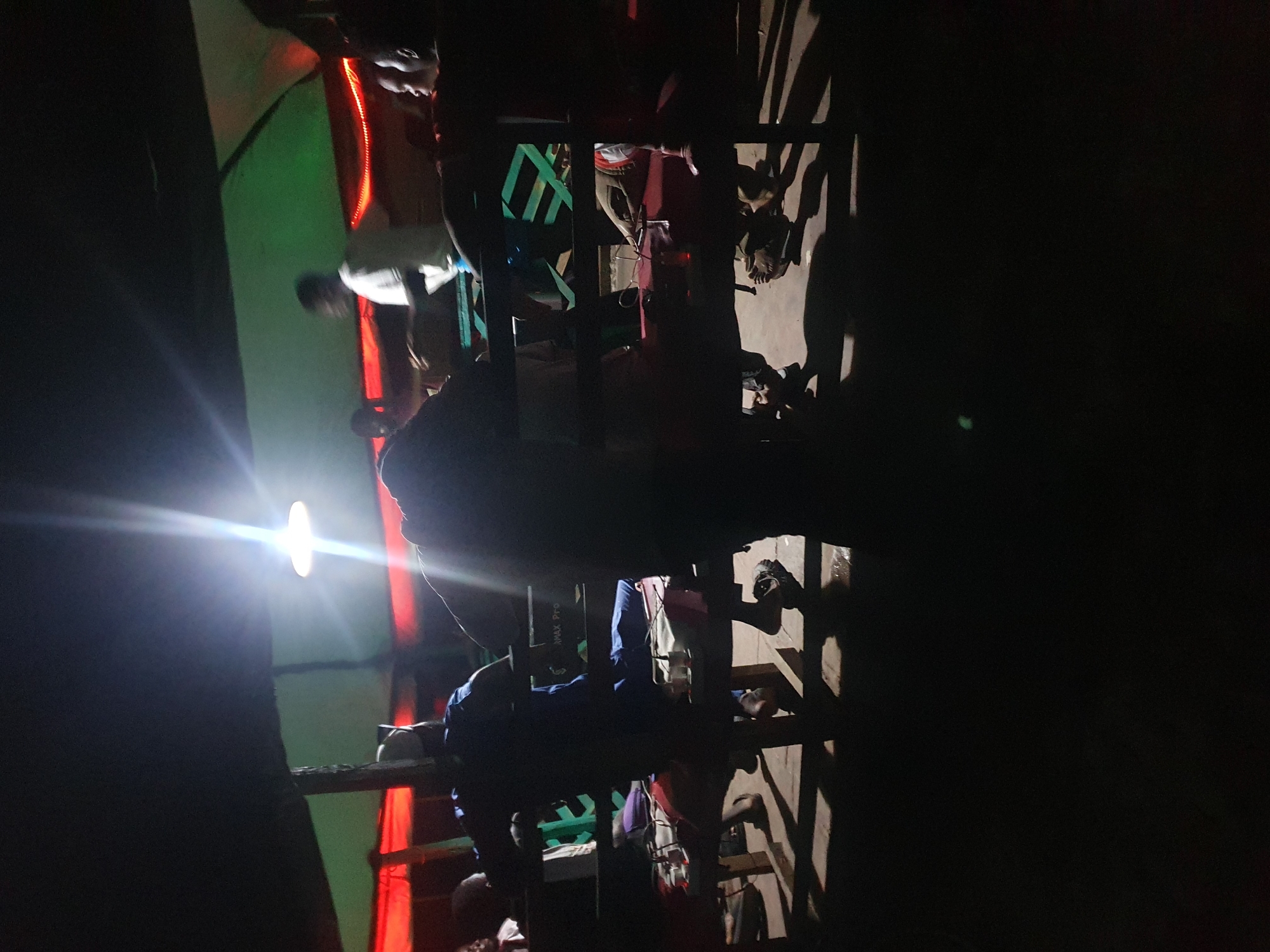 The image depicts a dimly lit outdoor gathering, likely at night, possibly in Lokoja, Nigeria. The scene appears to be under a makeshift roof structure supported by wooden poles. A bright overhead light casts harsh shadows and creates lens flares, obscuring some details. Several individuals are visible, silhouetted against the light or in the shadows. They appear to be seated or standing around tables or benches. The tables are covered with what look like red cloths and some objects are placed on them, possibly food or electronic devices. The overall atmosphere suggests a casual social event or a gathering at a roadside eatery. The background shows a green and red fabric, possibly part of the shelter or decorations, with red string lights. The ground appears to be concrete.