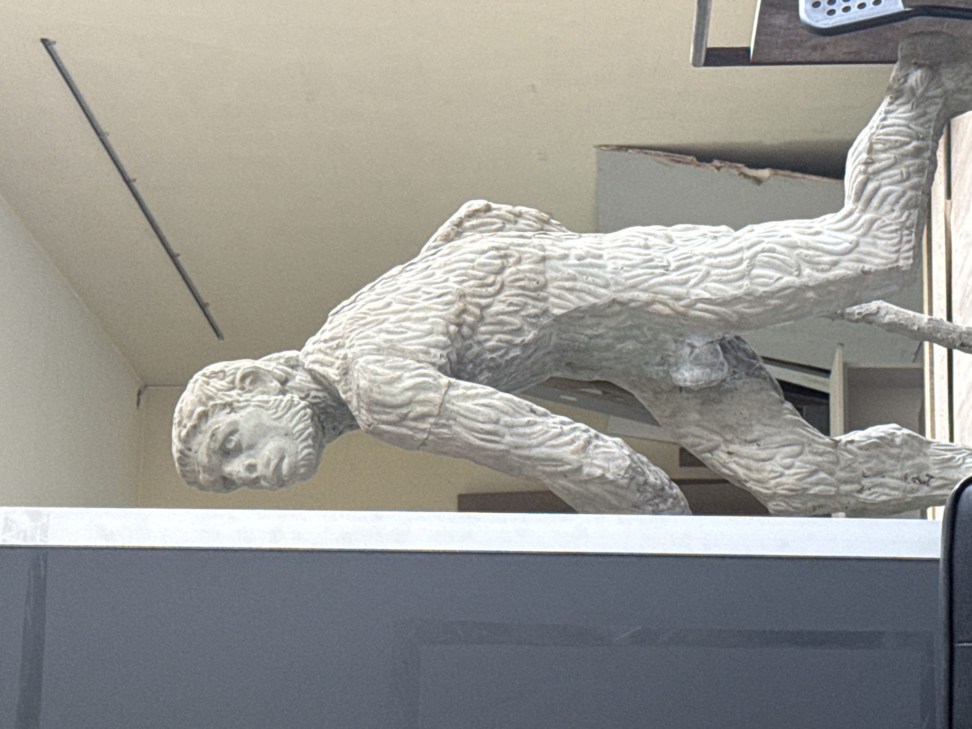The image shows a statue of a male figure on a building facade in Plovdiv, Bulgaria. The statue is light gray in color and appears to be made of stone or a similar material. The figure is depicted in a dynamic pose, leaning forward as if crawling or climbing. The statue has a weathered texture, with detailed striations across its surface. It is positioned above a modern building with a gray, reflective exterior. The background includes parts of the building's facade, with visible windows and architectural details. Above the statue, a beige-colored exterior wall is visible, with some architectural elements.