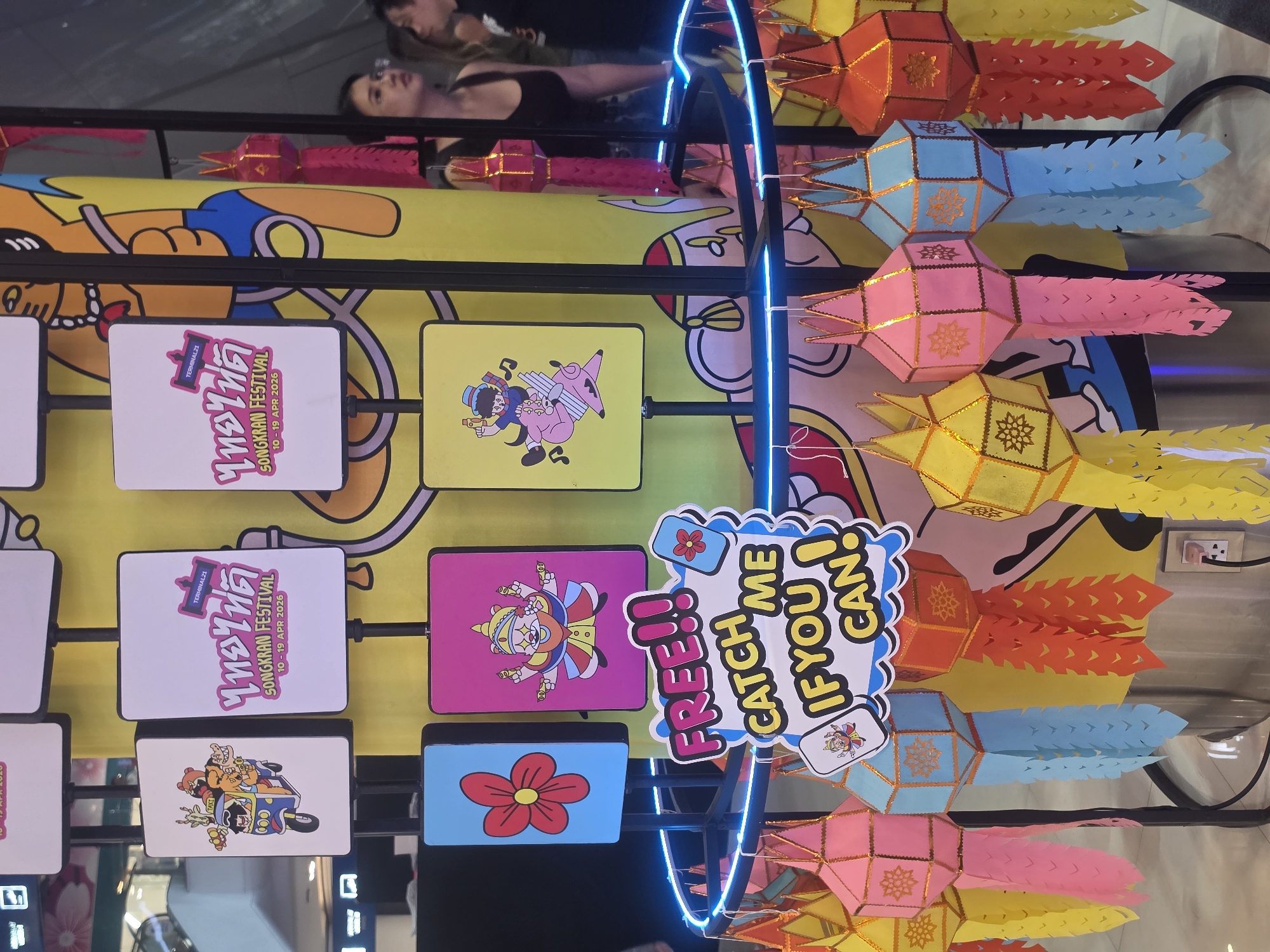 This image captures a vibrant and festive indoor display, likely within a shopping mall in Bangkok, Thailand, given the visible "TERMINAL21" branding. The scene is dominated by colorful paper lanterns, numerous illustrated signs, and a curved blue neon light structure.
Several strings of multi-colored lanterns, primarily red, light blue, pink, and yellow, hang horizontally across the right side of the frame. These lanterns are octagonal with intricate gold patterns and extended, cut-paper tails, typical of traditional Lanna style.
The main subjects in the center and left are a series of rectangular display panels. Two prominent white signs feature the text "TERMINAL21" and "SONGKRAN FESTIVAL 10 - 19 APR 2026" in English, alongside Thai script. Other signs display various cartoon-like illustrations, including a character riding an animal, a character with multiple arms, and a simple red flower.
Near the center-right, a playful speech bubble sign stands out, reading "FREE!! CATCH ME IF YOU CAN!" in large, yellow and blue letters, suggesting an interactive game or giveaway.
In the upper portion of the image, the reflections of several people, including a man and a woman, are visible on a glossy or mirrored surface, indicating an active public environment. The bright colors, festive decorations, and explicit mention of the "Songkran Festival" (Thai New Year) convey a lively and celebratory atmosphere.