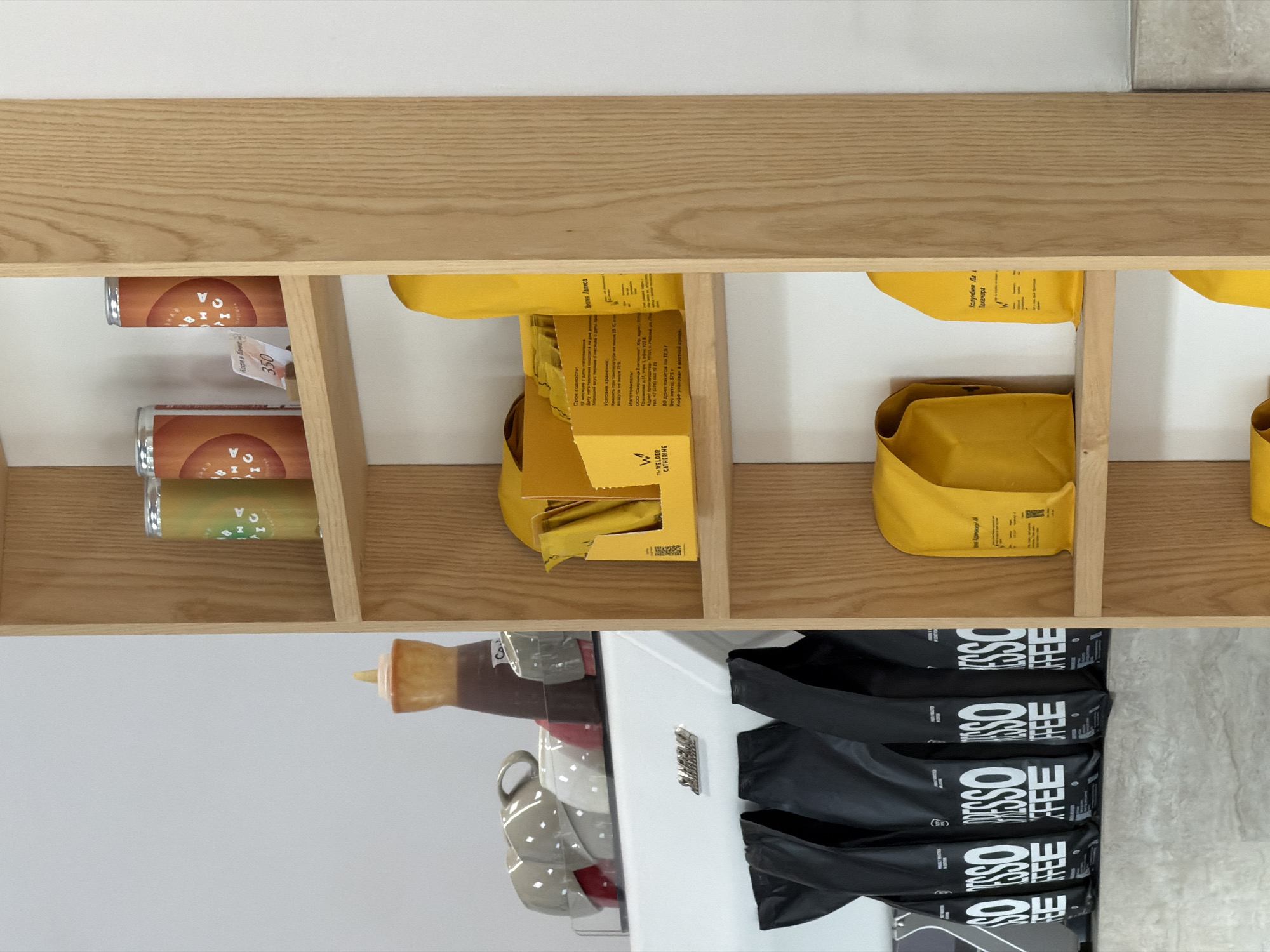 This eye-level shot presents a well-organized wooden shelf divided into several compartments. Each compartment contains various items, ranging from canned goods and yellow packages to black, branded coffee bags.
The leftmost section of the shelf holds several cans, varying in color from orange to green. Adjacent to these, in the middle compartments, are yellow packages, some of which appear to be partially opened, revealing their contents. A label on one of the packages reads "The WELDER CATHERINE" in addition to "Кофе в паке." There is additional Russian text that include "Срок годности", "Условия хранения" and "Изготовитель", which translate to "shelf life", "storage conditions", and "manufacturer" respectively.
The rightmost section of the shelf features several black coffee bags. Text on the bags reads "ESSO COFFEE" vertically, indicating the brand of coffee.
Below the shelf, a bottle and a stack of mugs can be seen, along with what appears to be a small refrigerator.
Without further information, it is not possible to pinpoint the specific location in Izhevsk, Russia.