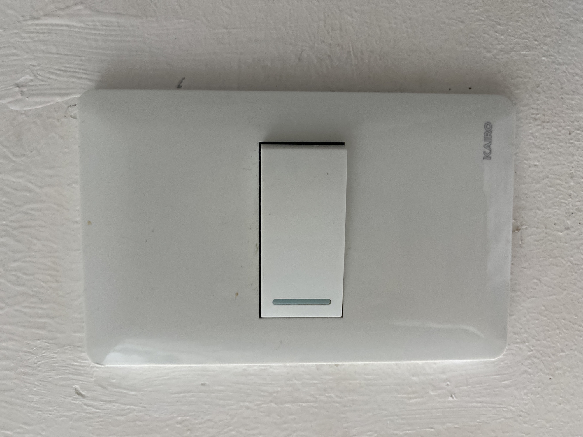 A white, single-gang electrical light switch, bearing the brand name "KAIRO" embossed in grey on its lower right corner, is centered on a textured, off-white painted wall. The switch features a rectangular, white rocker-style button, currently positioned to the left. A thin, vertical, translucent blue-green indicator strip is visible on the left edge of the rocker switch. The white plastic faceplate of the switch has rounded corners and a slightly glossy finish. The surrounding wall exhibits a coarse, troweled texture with minor scuffs and dirt specks. There are no discernible architectural or environmental features within the image to specify a precise location within Davao City, Philippines.
