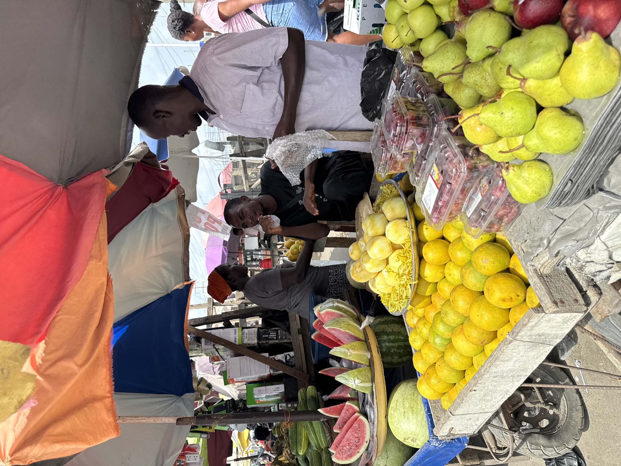 The image shows a fruit vendor in Lekki, Nigeria. A variety of fruits are on display including pears, grapes, lemons, watermelons, cucumbers, and more. The vendor, a man with an orange head covering, is standing behind the fruit stand. Two customers are present: one man in a white shirt looks at the fruit, while another in a black shirt appears to be eating something. A canopy made of various colored fabrics covers the fruit stand. The scene is a busy outdoor market.