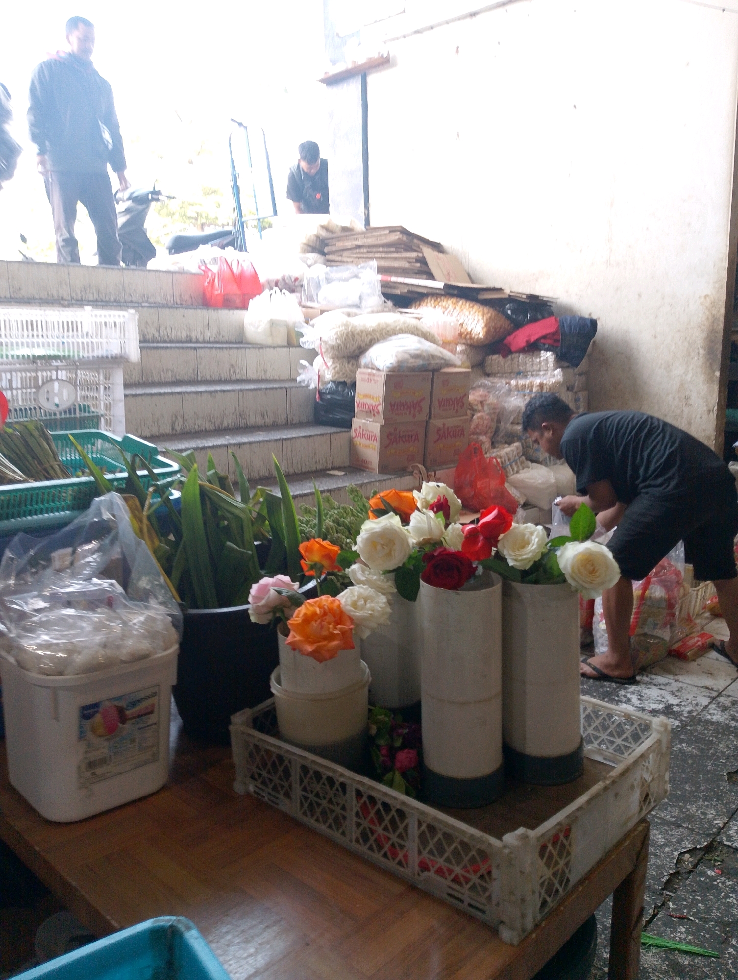 The image depicts an outdoor market stall in Lembang, Indonesia. A man in a black t-shirt and shorts is seen bending over to the right, appearing to arrange flowers. In front of him, on a wooden surface, are several arrangements of artificial flowers, primarily in shades of orange, white, and red, placed in white cylindrical vases. These vases are contained within a white plastic crate with a diamond pattern. To the left of the flowers, a large white plastic tub with a lid is visible, containing what appears to be food items. Further to the left, a green plastic basket filled with long, leafy green plants is present. Behind the flower stall, a set of concrete steps leads upwards, with a person in a black jacket and pants standing at the top. Another person, possibly a vendor, is visible behind a counter or display at the top of the steps. The background shows stacked boxes, bags of goods, and a motorcycle parked on the steps, suggesting a busy market environment. The ground is a mixture of concrete and dirt.