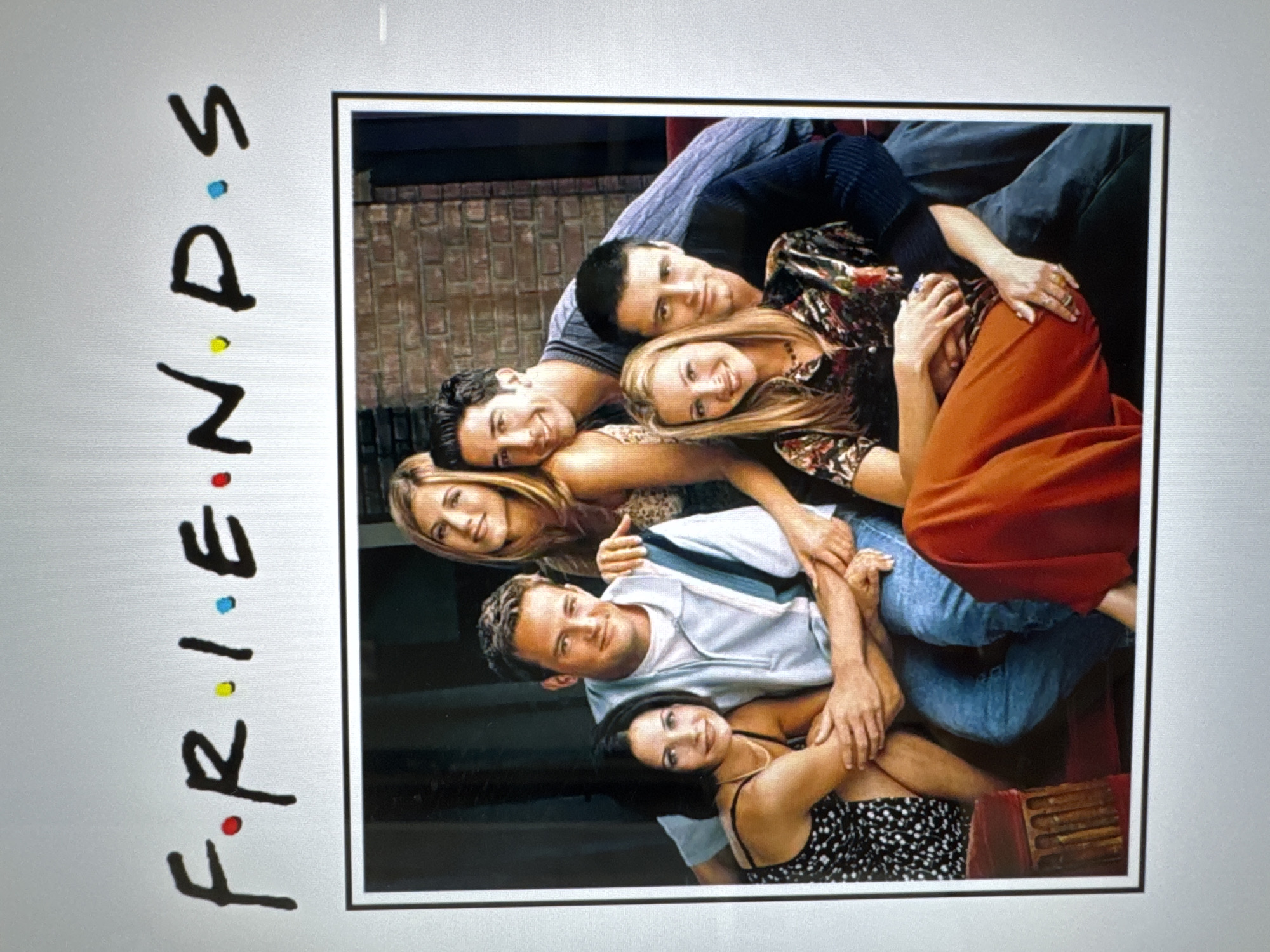 The image is a promotional poster for the television show "Friends". The cast is arranged in a close, overlapping group portrait. From top to bottom: Joey, Phoebe, Ross, Rachel, Chandler, and Monica. The background is a brick wall. The title "F•R•I•E•N•D•S" is vertically aligned on the left, with colored dots in place of periods. The image's setting is not specified.