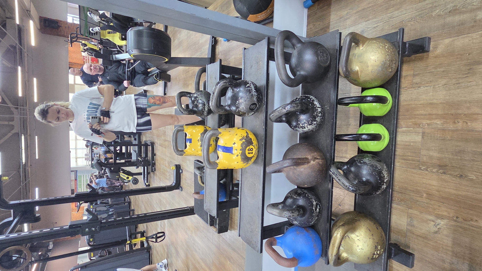 The image captures an indoor scene within a brightly lit gym or fitness center, featuring a woman taking a mirror selfie among various exercise equipment.
In the foreground, a blonde woman, with her hair tied back, is the primary subject. She wears a white t-shirt with "BROOKLYN" and "1908" (reversed from reflection) printed on it, patterned shorts, and black fingerless workout gloves. She holds a smartphone to take the picture, her expression neutral. A prominent tattoo of a woman's face, possibly with blue hair or headwear, is visible on her left thigh/hip.
To the woman's right, a multi-tiered metal rack holds an array of kettlebells. These include black, gold/bronze, and yellow kettlebells, with one yellow one clearly marked "18." Two neon green kettlebells with black handles are also visible on the top shelf.
The gym features a light wooden floor and light-colored walls, with sections of exposed red brick in the background. Industrial-style lighting fixtures hang from the ceiling. In the background, reflected in the mirror or glass, several other individuals are present. Two men are visible, one bald, wearing dark athletic wear and seated on a gym machine, and another partially obscured. Further back, more gym-goers are exercising on various weight machines and treadmills. The overall atmosphere suggests a typical daytime workout session in Chaikovsky, Russia.