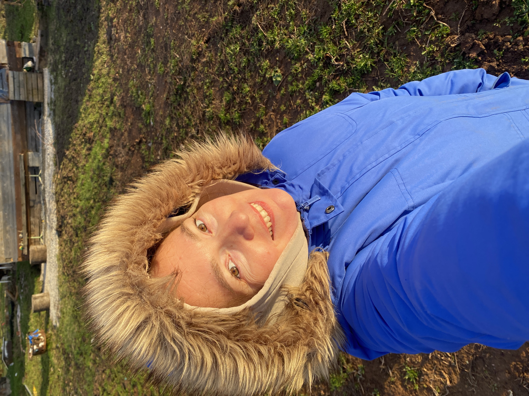 The central subject of this image is a person, likely female, captured in a close-up selfie outdoors. She is looking upwards towards the camera, beaming with a wide, cheerful smile that reveals her teeth. Her eyes are brown. She is dressed in a vibrant blue winter jacket or parka featuring a voluminous, light brown faux-fur trim around the hood. A light-colored fabric band is visible around her neck or chin, beneath the hood. The brand name "Volcom" is faintly discernable on a zipper pull near her collar.
The setting is an outdoor, natural environment, illuminated by bright daylight which casts clear shadows and highlights the textures of the fur and her skin. The background consists of rough, earthy ground sparsely covered with patches of green grass and dry vegetation. In the left portion of the image, some rustic wooden structures, possibly parts of a fence or a small building, are visible, suggesting a rural or countryside location. The person's expression conveys happiness and contentment. This scene is located in Ольховка, Russia.