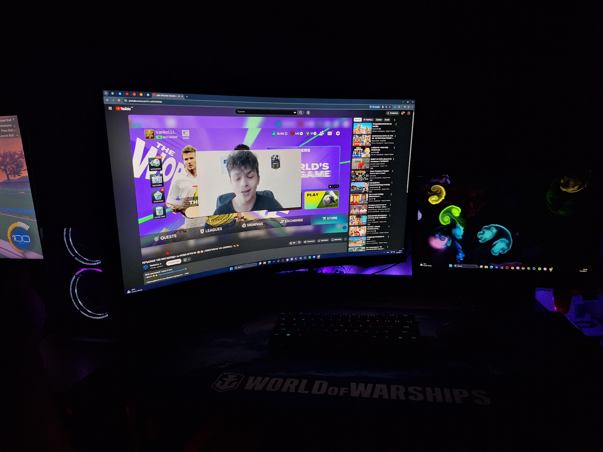 The image shows a dark room with a gaming setup. A large, curved monitor displays a YouTube page with a gaming video playing. To the right of the main monitor, a second smaller monitor shows a colorful, abstract wallpaper. In front of the monitors, a keyboard with illuminated keys is visible. The overall lighting is dim, with purple and blue LED lights from the computer tower and the monitor illuminating the scene. A large mousepad with the "World of Warships" logo is placed on the desk. The scene appears to be a personal gaming space, likely in a residential setting. The specific location in Varna, Bulgaria cannot be determined from the image.