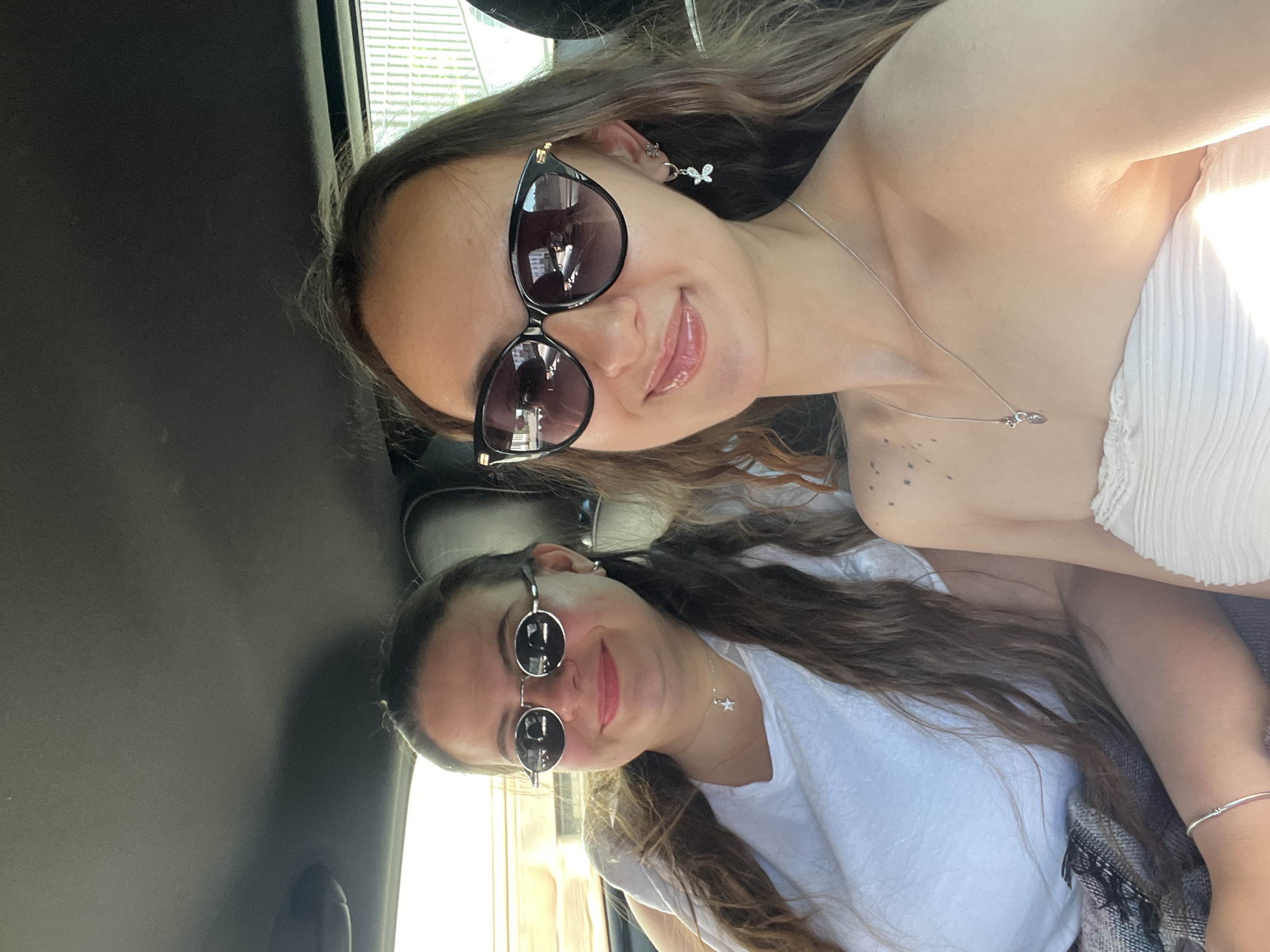 The image is a selfie taken inside a car, rotated 90 degrees counter-clockwise. Two young women are visible, presumably the occupants of the vehicle.
The woman in the upper part of the image wears large, dark-framed sunglasses, butterfly earrings, and a silver necklace. She has long, wavy brown hair. Her top appears to be white. Visible on her shoulder are a cluster of small star-shaped tattoos.
The woman in the lower part of the image also has long, wavy brown hair and is wearing sunglasses. Her sunglasses have a circular lens and a wire frame. She wears a white t-shirt and a silver necklace with a star pendant. She has a silver bracelet on her left wrist.
The interior of the car is mostly dark. The window is visible on the left side of the image. The precise location within Saarbrücken, Germany is not determinable from the given information.