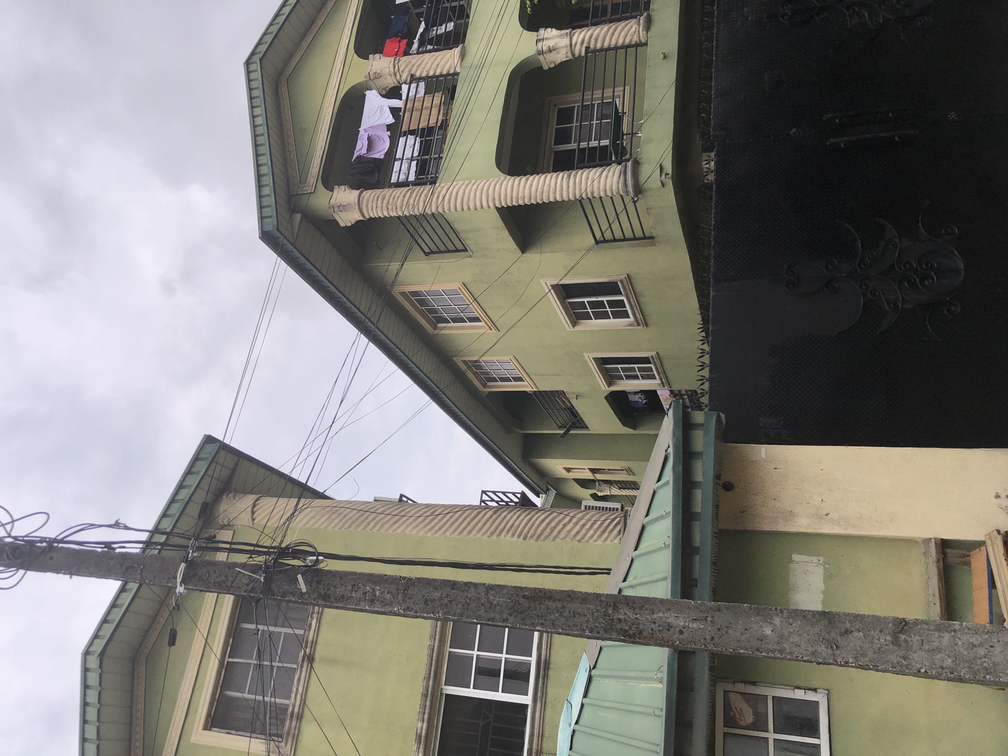The image depicts an overcast day in Ado, Nigeria, with two adjacent green buildings dominating the frame. A prominent utility pole, laden with numerous black wires, stands in the foreground to the left. The building on the left features a green corrugated metal roof overhang, an upper floor with white-framed windows, and decorative ridged architectural elements on its exterior. The building on the right is taller and has a more elaborate design, including arched balconies with black metal railings. Several items of clothing, including a white shirt and a purple garment, are hung out to dry on the balcony. The ground level of the right building has a window and a small table visible on the balcony. A substantial black metal gate with intricate scrolling patterns is positioned in front of the buildings, obscuring the lower portion of the scene. The sky is filled with gray clouds.