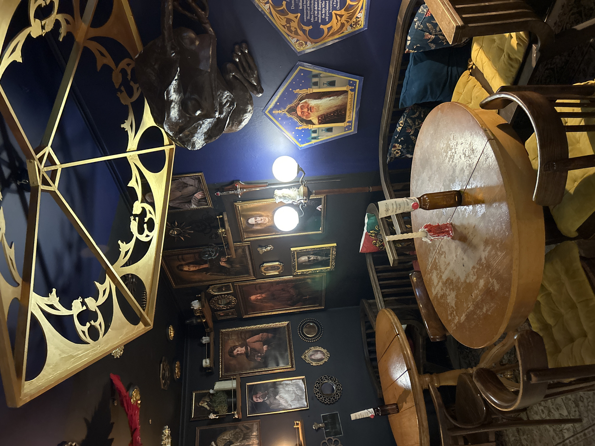 The image depicts a dimly lit room with dark blue walls, adorned with numerous framed portraits in ornate gold frames. A large, decorative gold metal structure hangs from the ceiling, featuring intricate scrollwork. On the walls, several portraits of individuals in historical attire are visible, along with decorative mirrors and a few shelves holding small objects. In the foreground, two round wooden tables are set with a bottle and a candle with melted wax on one, and a single bottle on the other. Wooden chairs with yellow cushions surround the tables. To the right, a wooden bench with plush cushions in floral and solid teal patterns is positioned against the wall, next to a framed image of Albus Dumbledore. A dark, sculpted object, resembling a gargoyle or creature, is mounted on the wall above the bench. The overall atmosphere suggests a themed establishment, possibly a cafe or restaurant, with a distinct wizarding or historical theme.