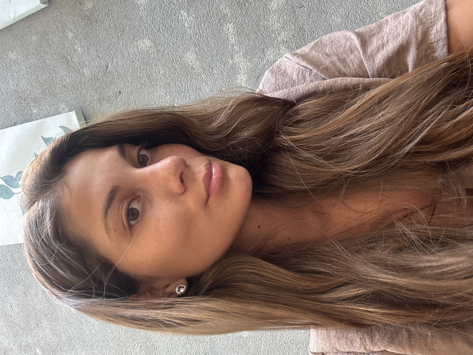 A woman with light brown hair and fair skin is facing the camera with her head tilted slightly. She has freckles and is wearing a stud earring in her left ear. She is wearing a light brown garment. The background is a textured gray surface and a light blue and white patterned object. The image does not provide any information about the location being Đà Nẵng, Vietnam, or any specific details to confirm that location.