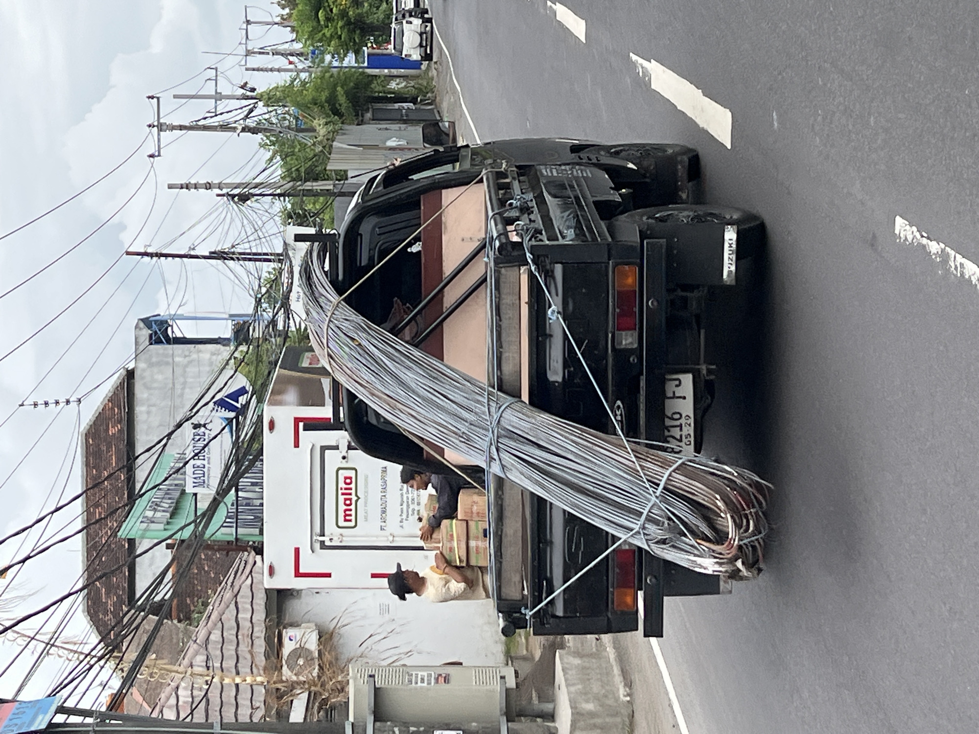 A black pickup truck with a large bundle of metal rods tied to its bed drives down a paved street in Denpasar, Indonesia. Two men, one wearing a light-colored shirt and a dark cap, the other in a dark shirt, are standing near the truck, engaged in stacking boxes. The truck bed contains some wooden planks and other materials beneath the metal rods. A white refrigerated truck with the logo "malia" is parked behind the pickup truck. Buildings with tiled roofs, some with signs indicating businesses, line the street. A complex network of electrical wires crisscrosses overhead, supported by utility poles. Other vehicles, including a white SUV, are visible further down the street. The sky is overcast.