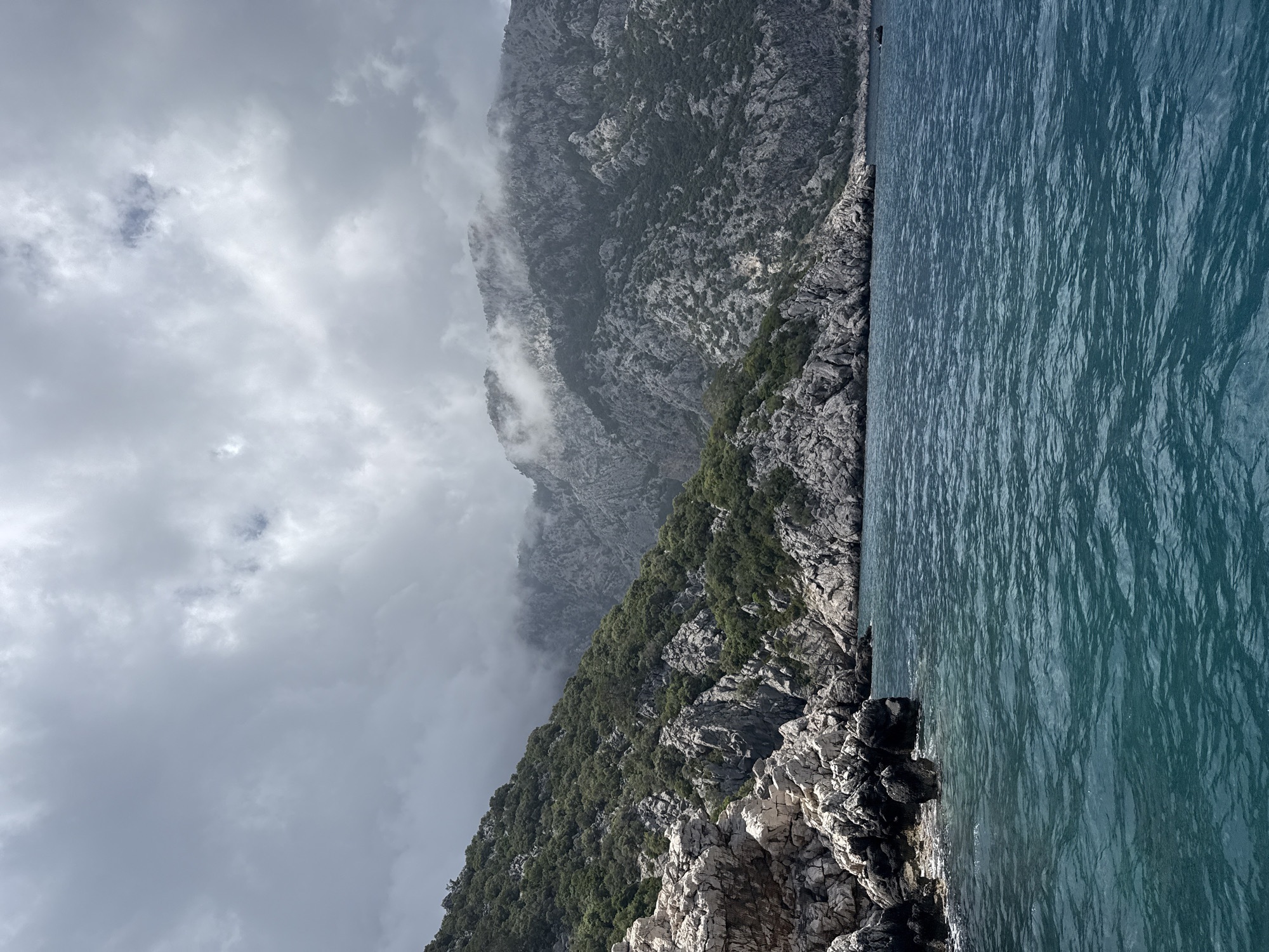 The image depicts a coastal landscape in Kumluca, Turkey. A mountain range, partially obscured by low-lying clouds, rises along the coastline. Vegetation covers parts of the mountainside. The sea stretches out, its surface ruffled by the wind. The foreground consists of rocky terrain.
