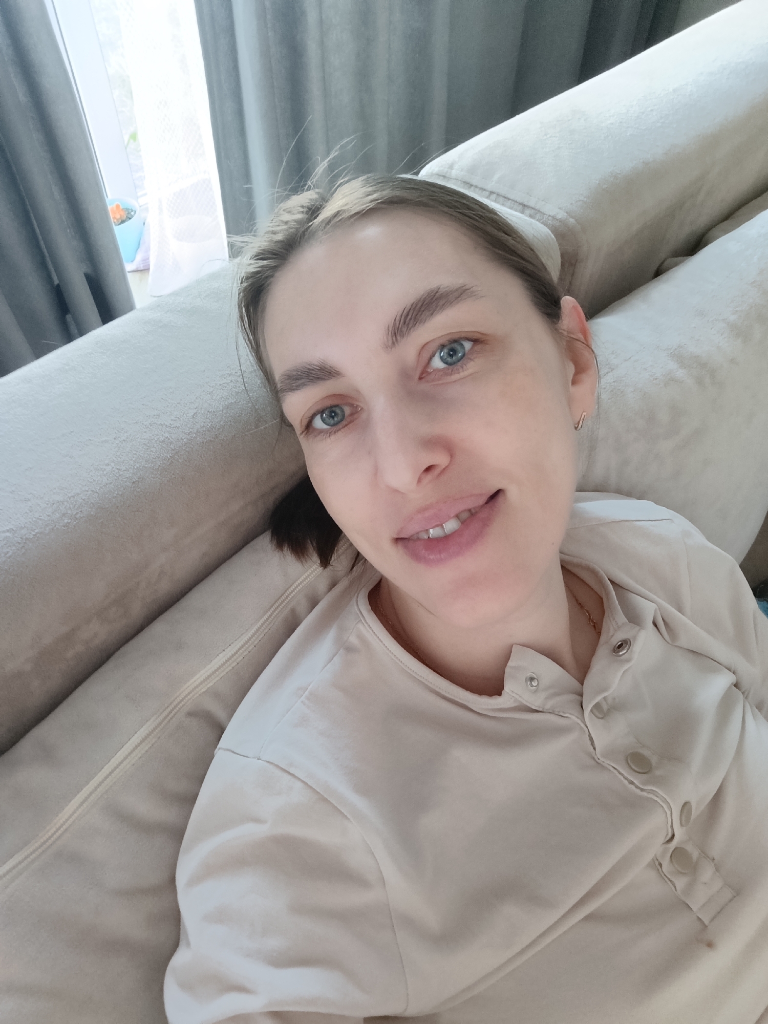A woman lies comfortably on a light-colored sofa, taking a selfie. She has fair skin, blue eyes, and light brown hair pulled back from her face, revealing prominent eyebrows. She is wearing a light beige or cream long-sleeved top with snap buttons down the front, and a thin gold chain is visible around her neck. A small gold hoop earring can be seen in her right ear. Her expression is relaxed and pleasant, with a gentle smile as she looks directly at the camera.
The sofa she is resting on is upholstered in a soft, light-colored fabric. In the background, to the left, a large window allows bright natural light to stream in, indicating it is daytime. Gray or beige curtains are partially drawn on either side of the window. A small plant in a light blue pot is visible on the windowsill. The overall scene suggests a calm and casual indoor setting. There is no visible text in the image.