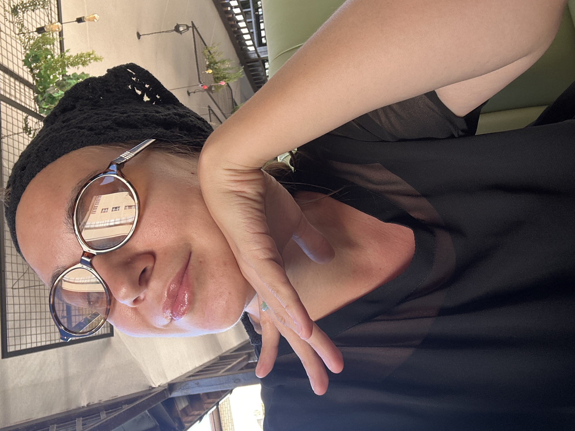 A woman wearing glasses and a black beanie is leaning on her hand, looking upwards and to the side. She is wearing a sheer black top. The background appears to be an outdoor cafe with hanging plants and lights. The scene is set in Vityazevo, Russia.