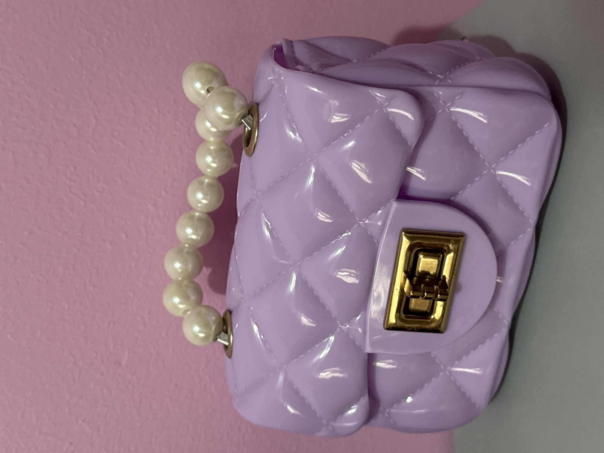 This is a close-up, vertical, eye-level shot of a small, quilted, lavender-colored purse with a pearl handle. The purse has a gold clasp on the front. It is positioned against a background that transitions from a light pink on the left to a light gray on the right. The lighting suggests an indoor setting, possibly a studio or well-lit room, with no specific time of day or weather indicated. The focus is entirely on the purse, highlighting its texture and sheen. There is no visible text in the image. The scene appears to be a still life, with the object isolated for presentation.