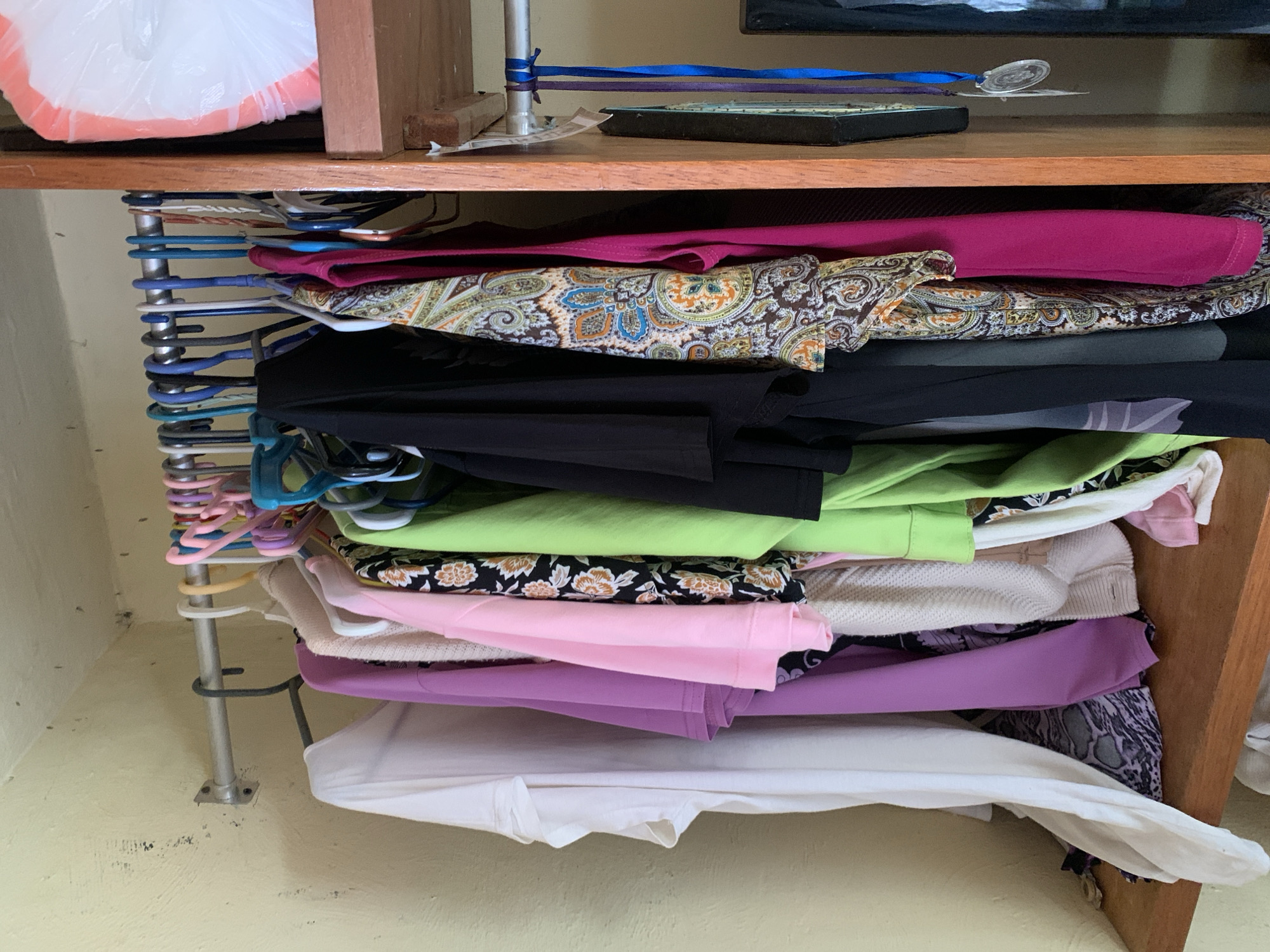 The image shows a closet filled with clothing. The closet has a wooden shelf and a metal rod where clothes are hung on hangers. There are various garments, including t-shirts, dresses, and what appear to be scarves. The background is a light yellow wall with some dark marks on it. On the right side of the image, there is a wooden frame and a white plastic bag with pink trim on a shelf. There are also some ribbons and a medal hanging from the rod. The scene is likely a domestic setting.