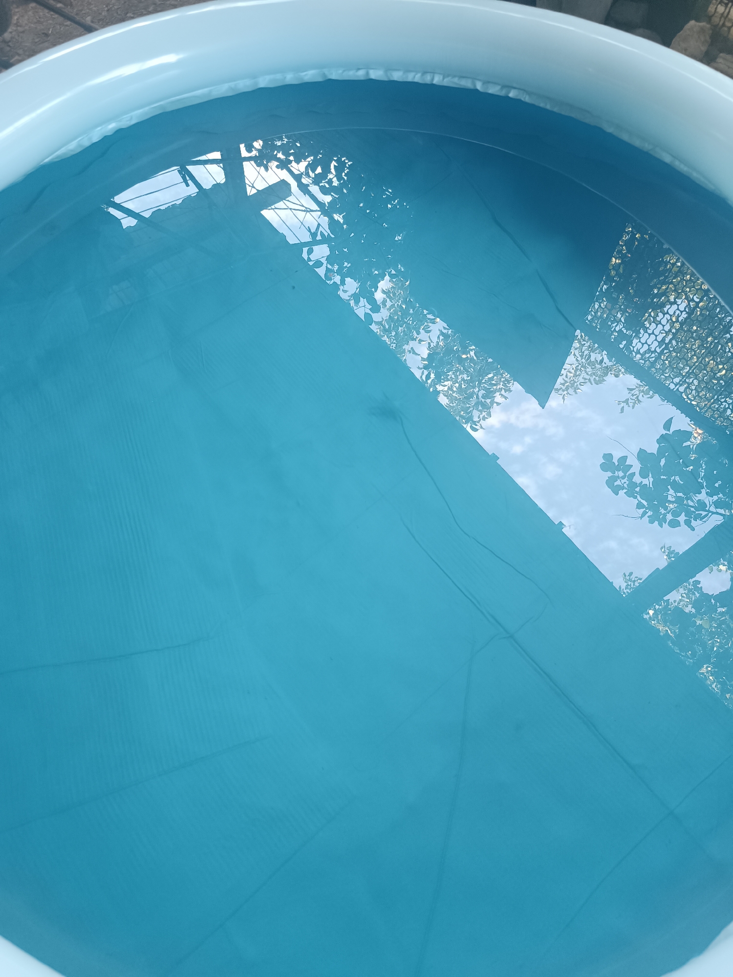 An overhead shot shows the interior of an inflatable pool filled with blue water. The water reflects the sky and some foliage, indicating an outdoor setting. The pool's white rim is visible at the top and bottom left edges of the frame. The water's surface shows some ripples and a texture from the pool liner beneath, with faint lines and indentations visible. The reflection in the water reveals a lattice structure with leaves and branches from trees, as well as a cloudy blue sky. The scene is static and depicts an empty pool.