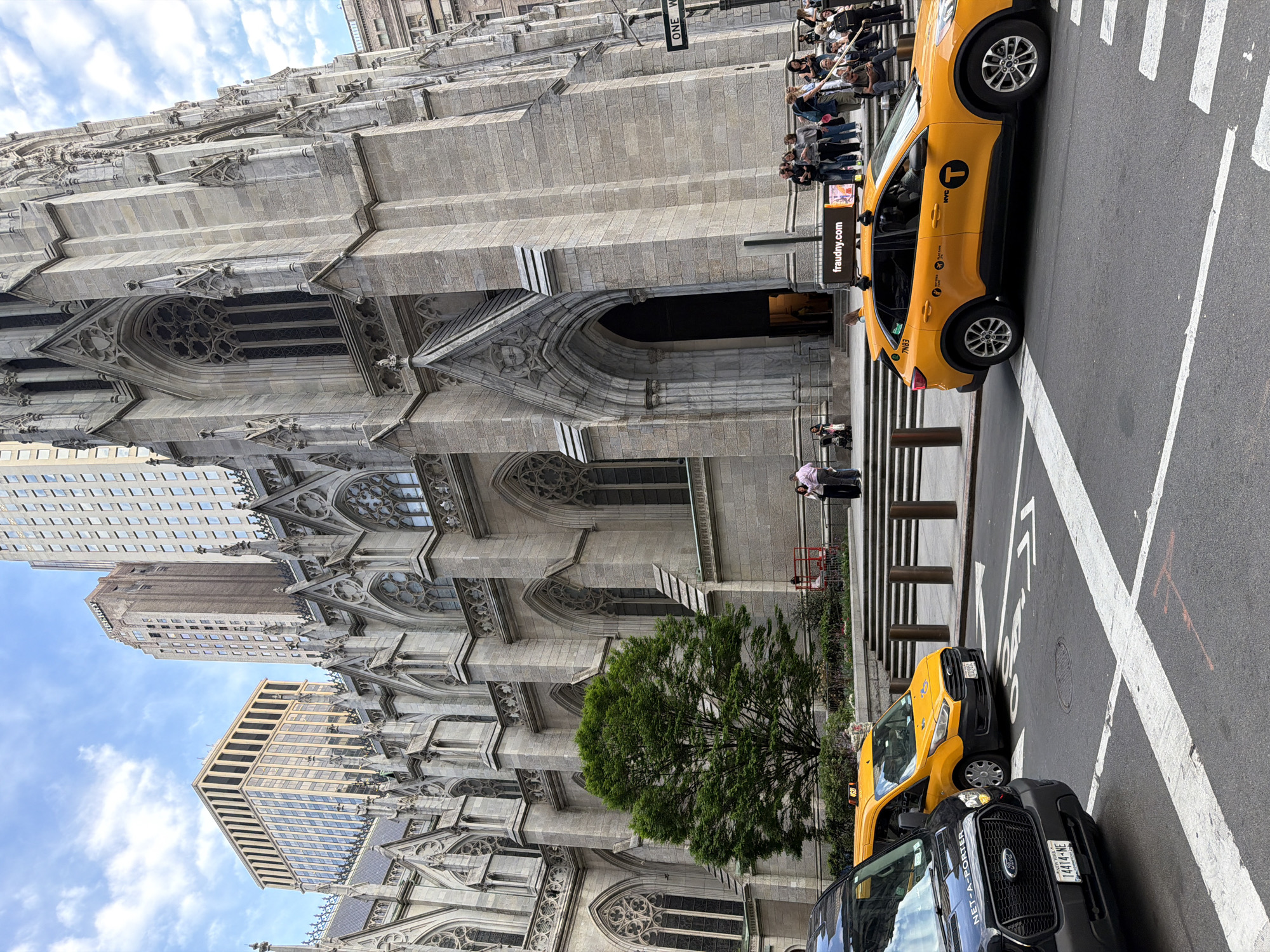 The image presents a street scene in New York City, United States. The architecture prominently features a large, historic cathedral. Adjacent to the cathedral, a busy street is visible. A line of people is standing by the cathedral. Several taxicabs are parked on the street, including one marked "NYC". A vehicle with license plate "14414-NE" is parked in front of one taxicab. An advertisement on a pole shows "fraudny.com". The sky is visible in the background with some clouds.