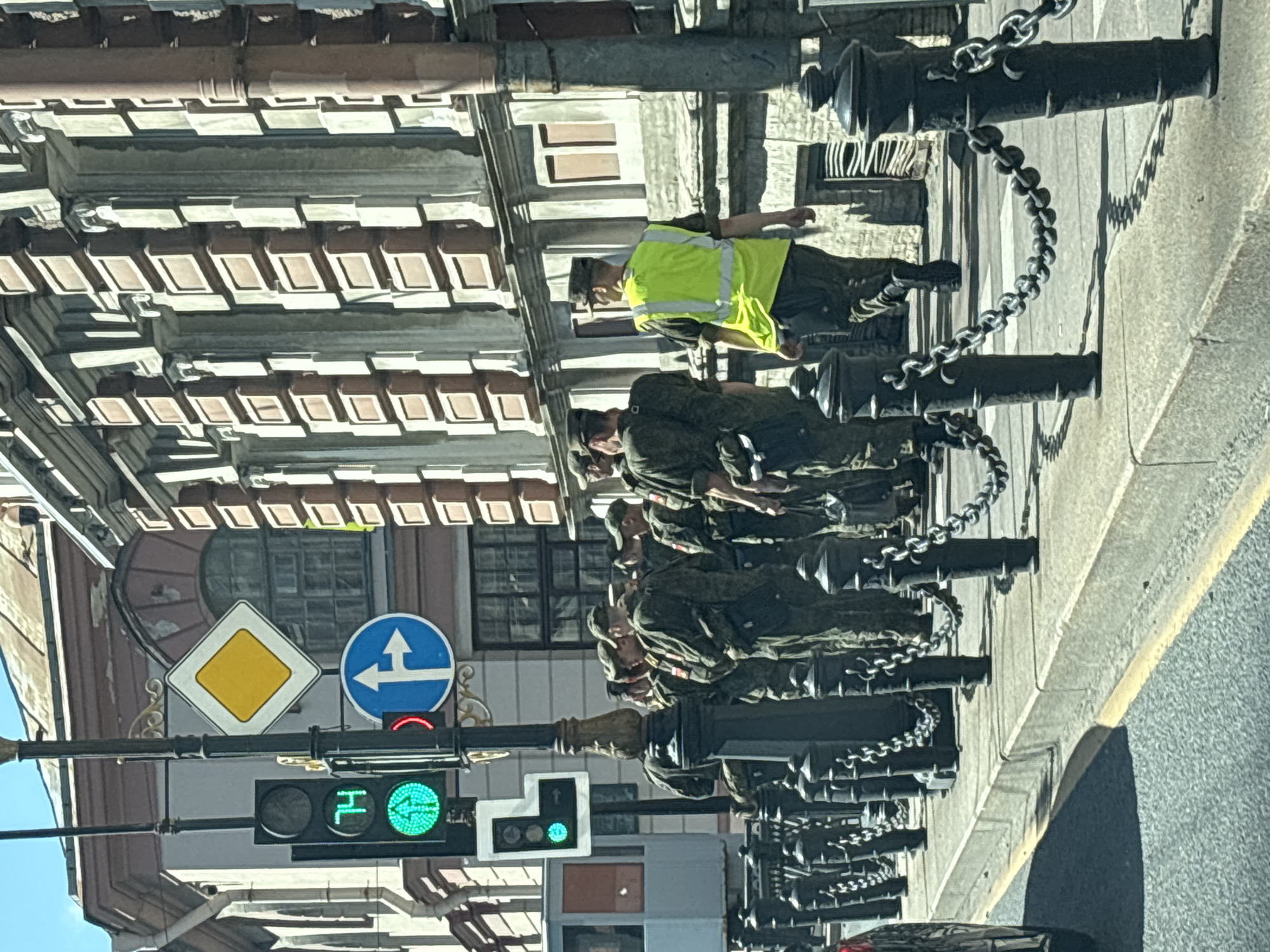 A group of men in military uniform stand on a sidewalk next to a building in Saint Petersburg, Russia. One man in the foreground wears a bright yellow safety vest over his uniform and walks away from the camera. The sidewalk is lined with black bollards connected by chains. A traffic light shows a green signal with an upward arrow. The building behind the men has a classical architectural style with columns and decorative elements. A street runs in front of the sidewalk.