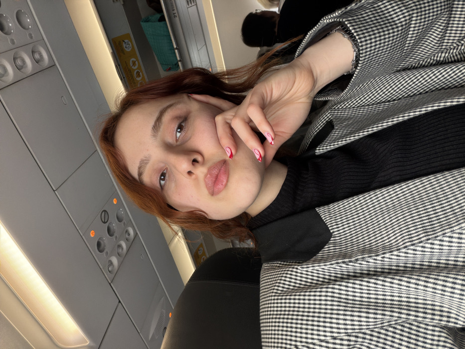 The image captures a female individual inside a commercial aircraft cabin. The scene is contextually placed in Tuzla, Turkey.
**People:**
A young adult female is centered in the frame, directly facing the camera in a self-portrait. She possesses light skin, reddish-brown hair, and dark eyes. Her lips are slightly puckered in what appears to be a "duck face" expression. Her left hand is raised, with fingers gently touching her chin and cheek. Her nails are painted light pink with distinct red wavy or heart-like designs. A silver-colored bracelet is visible on her right wrist. In the background on the far right, the top of another passenger's head, with dark hair, and shoulder are partially visible, facing away from the camera.
**Subjects/Objects:**
The primary subject is the female individual. She is wearing a black, ribbed turtleneck top under a black and white small-checkered blazer. The blazer features solid black lapels.
The immediate environment is a commercial airplane interior. Above the subject's head, the gray overhead passenger service unit is visible, equipped with individual reading lights, air vents, and call buttons; some indicator lights appear orange. Overhead bin doors are present, one with a yellow safety sticker and another containing a teal-colored bag. An illuminated strip light runs along the cabin ceiling. A black airplane seat headrest is partially visible on the left.
**Actions/Interactions:**
The female individual is engaging in a self-portrait, holding her left hand to her face in a casual pose. No direct interaction with other individuals is depicted.
**Environment:**
The environment is an airplane cabin, characterized by standard overhead panels, lighting, and seating arrangements of a commercial airliner. The interior appears clean and well-lit by both cabin lighting and an ambient glow from an unseen source (possibly a window or additional cabin lights).