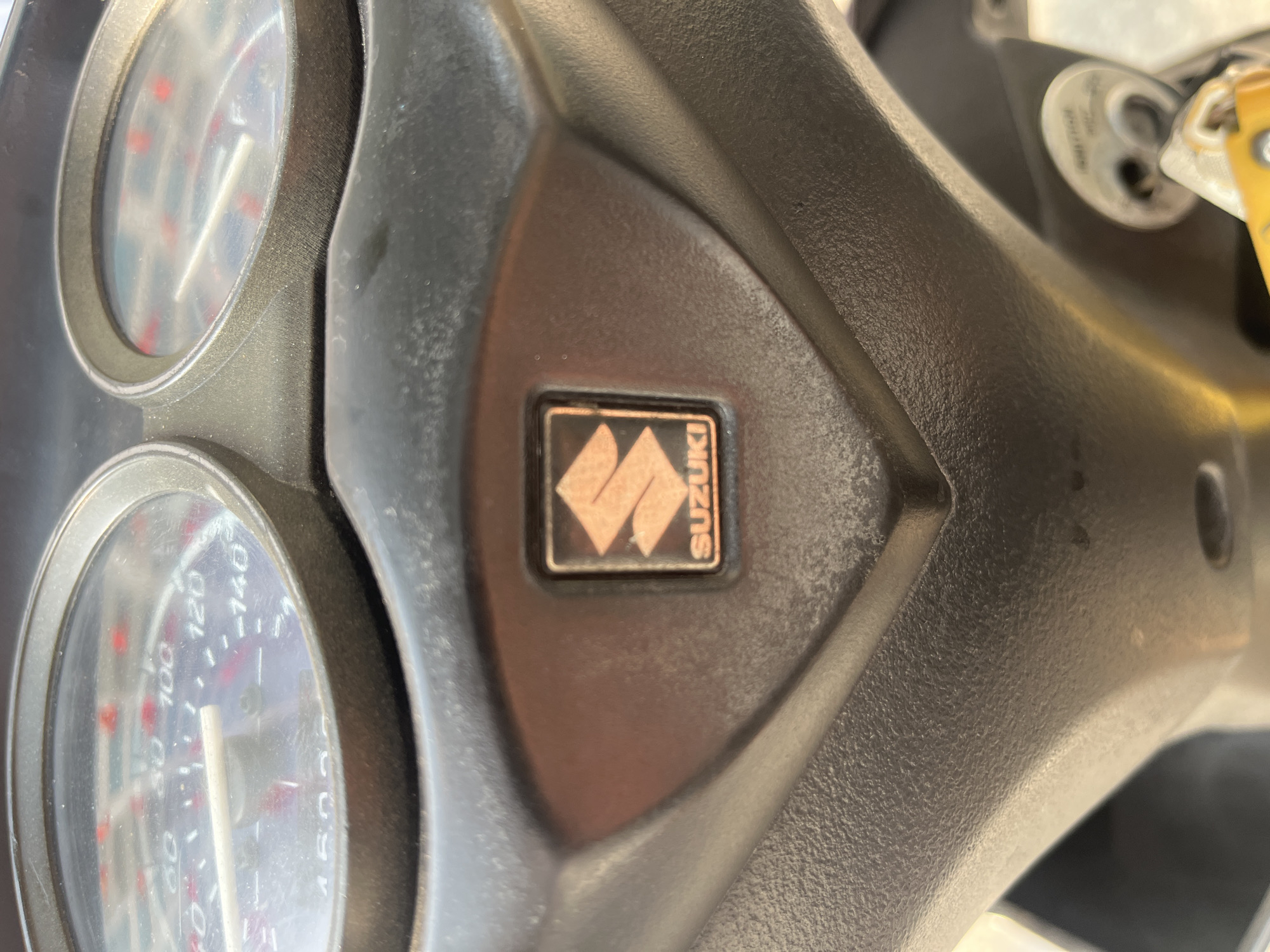 The image shows the dashboard of a Suzuki scooter. The dashboard features two circular gauges and a central panel displaying the Suzuki logo. A key is visible in the ignition switch. The location is assumed to be Bắc Nha Trang, Vietnam, but there is no visible evidence to confirm this.