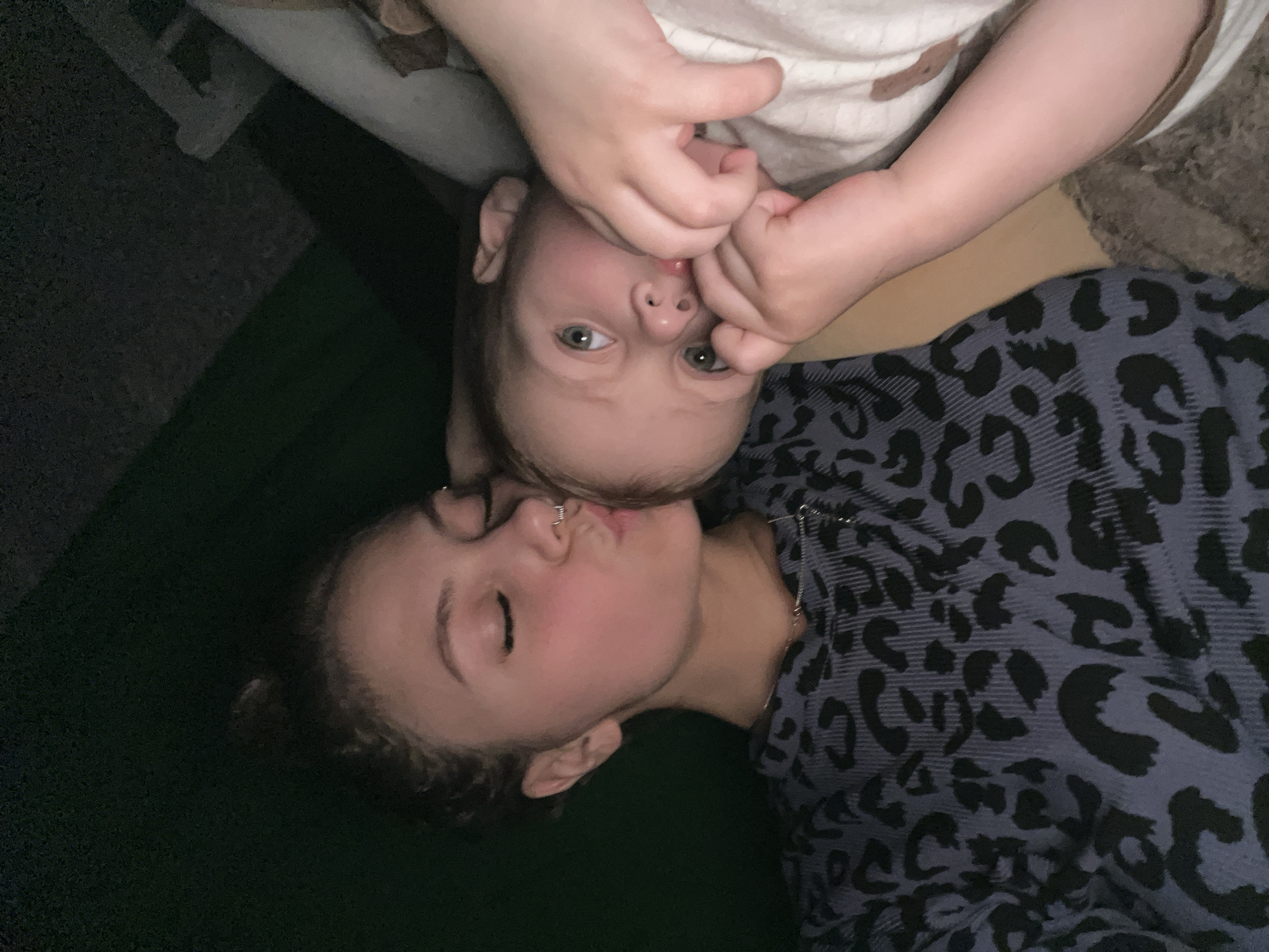 A woman and a toddler are lying down together, angled to the left. The woman has curly brown hair pulled back from her face and a nose ring, her eyes are closed and her lips pursed in a kiss. She is wearing a blue and black leopard-print shirt. The toddler is above her, with chubby hands over their mouth. Their eyes are open and looking at the camera. They are wearing a light-colored shirt. The background is dark. The image appears to have been taken indoors. It is not possible to determine the location of this image as Фоки, Russia.