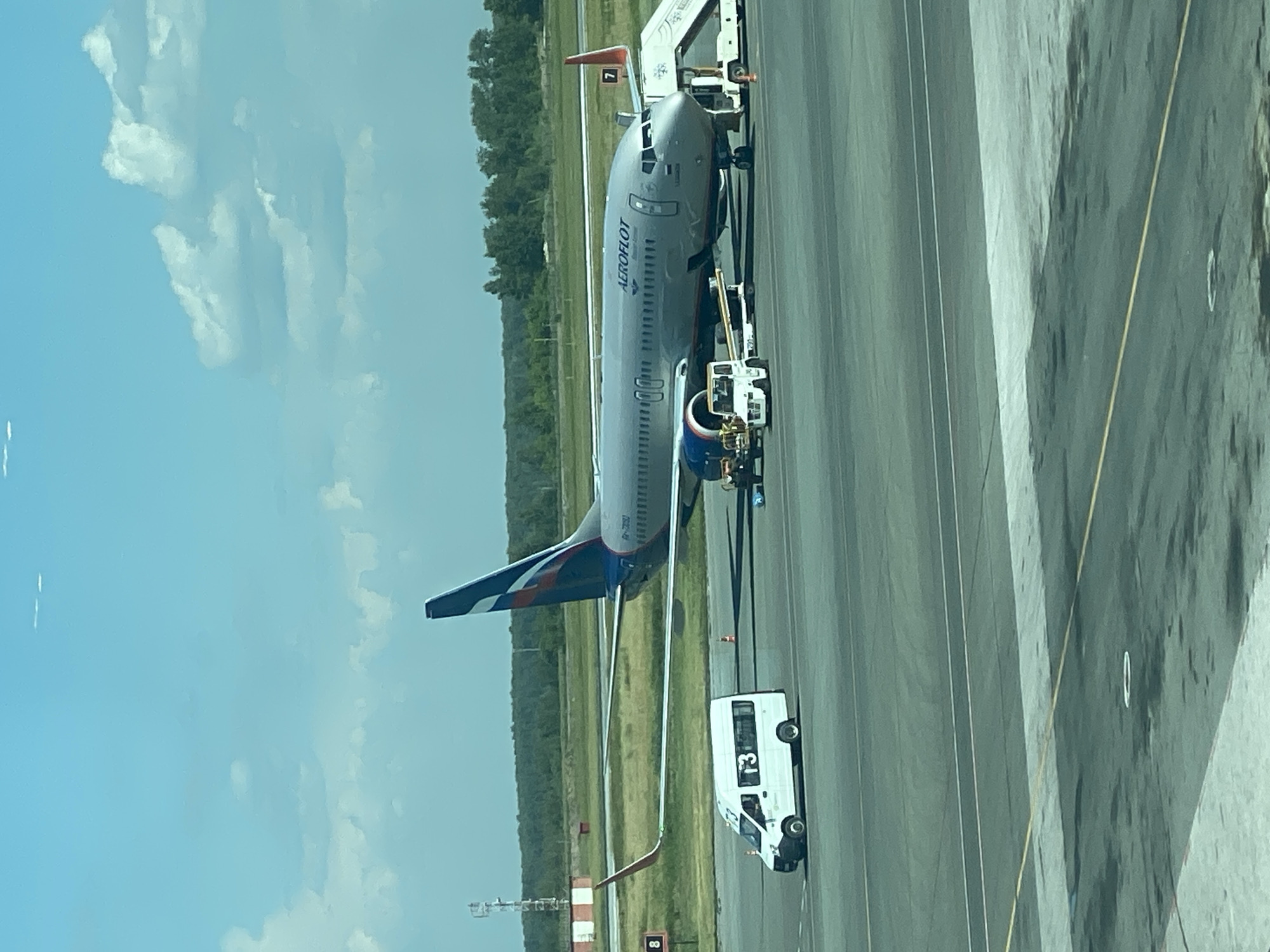 A wide-angle, eye-level shot captures an Aeroflot airplane parked on the tarmac at Tolmachevo Airport (IATA: OBT, ICAO: UNBB) in Ob, Russia. The aircraft, identified by the "AEROFLOT" logo on its fuselage and a Russian flag on its tail, is a medium-haul passenger jet, likely a Boeing 737 or Airbus A320 family aircraft, with its engines visible. A set of mobile stairs is positioned at the front entrance of the aircraft, and several ground support vehicles are stationed near the plane, including a white shuttle bus with "T3" labeled on its side, a baggage tractor, and a tow vehicle.
The scene is set against a bright, clear sky with scattered white clouds. In the background, a line of green trees forms a backdrop, and a communication tower with red and white markings is visible to the left of the frame. The tarmac is marked with various painted lines and a few traffic cones are scattered around the area. The overall impression is of a busy airport environment on a sunny day. No people are clearly visible in the image.